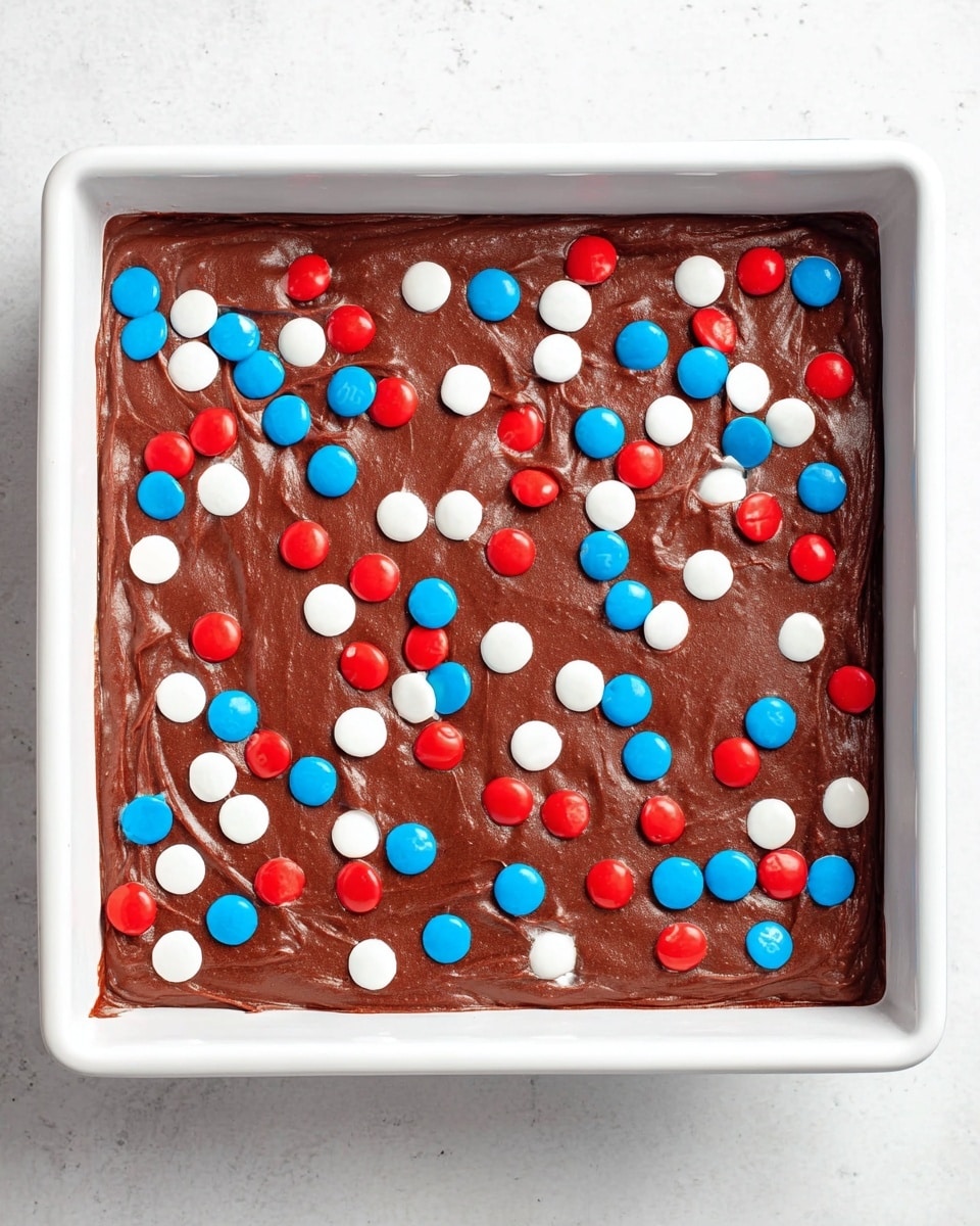 A square white baking pan is filled with one thick layer of smooth, rich brown chocolate batter, evenly spread across the pan. The chocolate layer is decorated with scattered small round candies in red, white, and blue colors, adding bright pops of color on the surface. The pan is placed on a white marbled surface, enhancing the contrast with the dark chocolate and vibrant candies. photo taken with an iphone --ar 4:5 --v 7