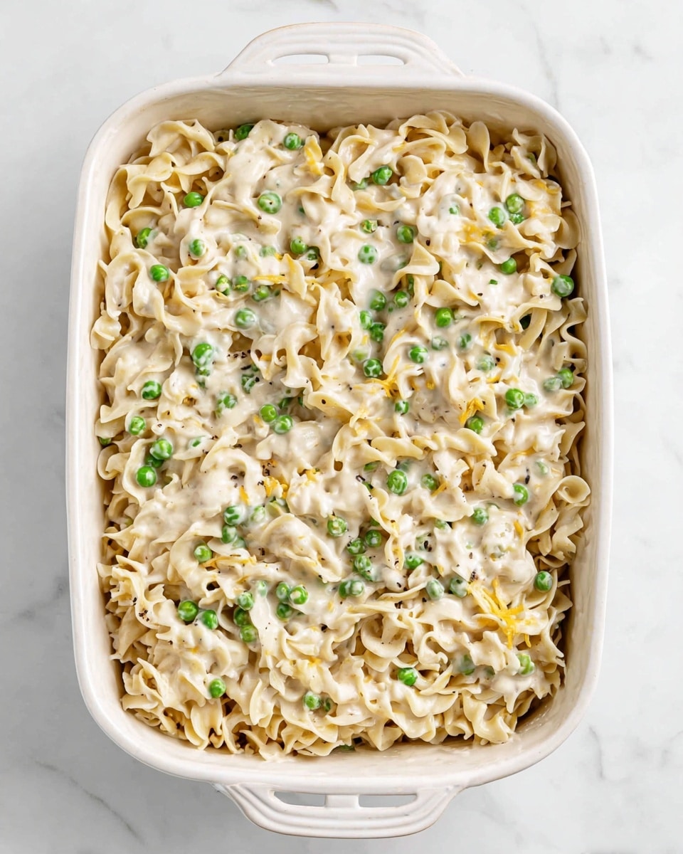 A white ceramic baking dish filled with a creamy casserole made of three layers: the base layer with a mix of soft, light beige egg noodles; the middle layer with small bright green peas evenly spread; and the top layer is coated with a smooth, creamy white sauce that slightly covers the noodles and peas, showing a soft texture with hints of melted yellow cheese peeking through. The dish is set on a white marbled surface. photo taken with an iphone --ar 4:5 --v 7
