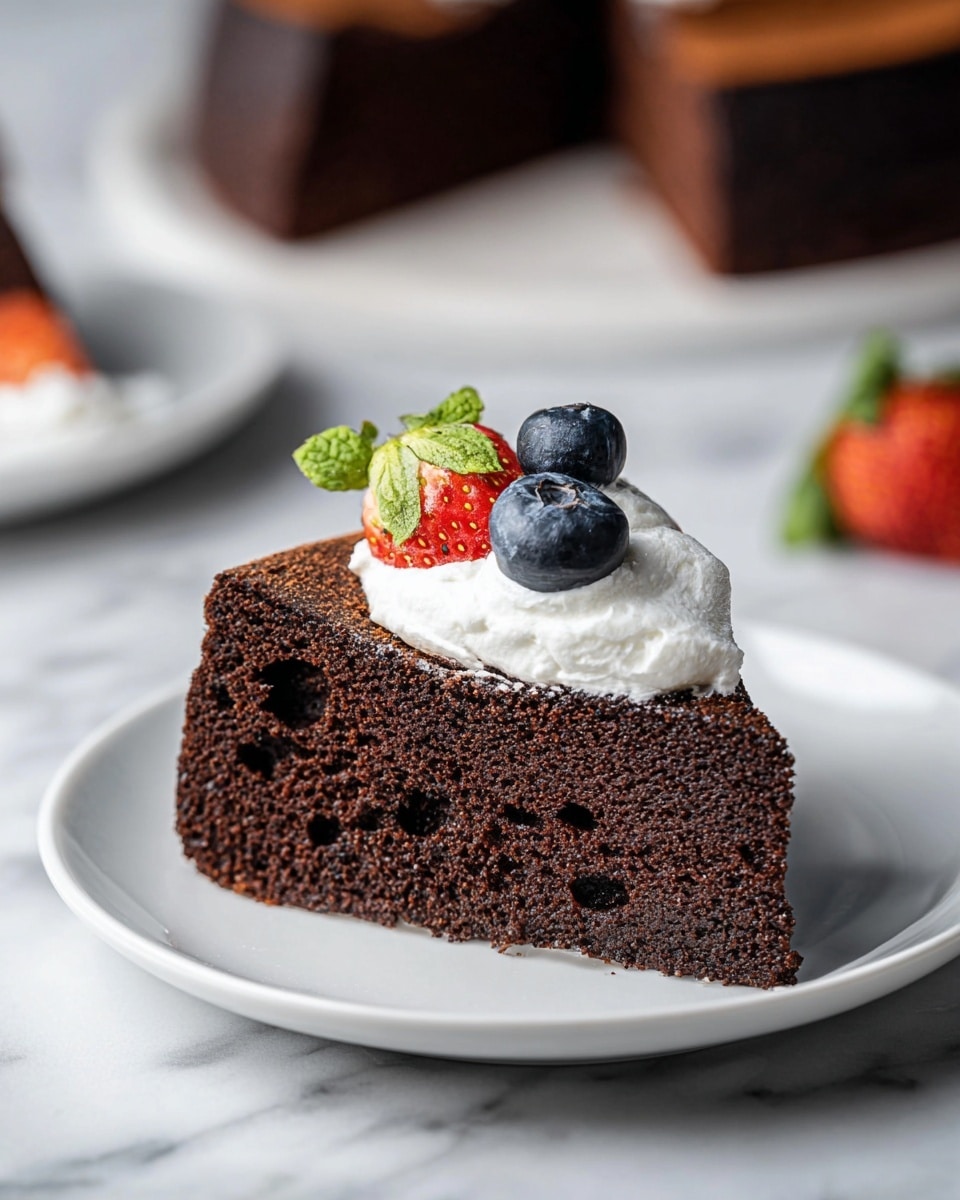 A slice of rich dark chocolate cake sits on a white plate, showing a moist and dense texture with small air holes inside. On top, there is a layer of light whipped cream, decorated with two fresh blueberries and a small piece of light red strawberry with its green leaf still attached. The cake and plate are placed on a white marbled surface, with another similar slice of cake blurred softly in the background. Photo taken with an iphone --ar 4:5 --v 7
