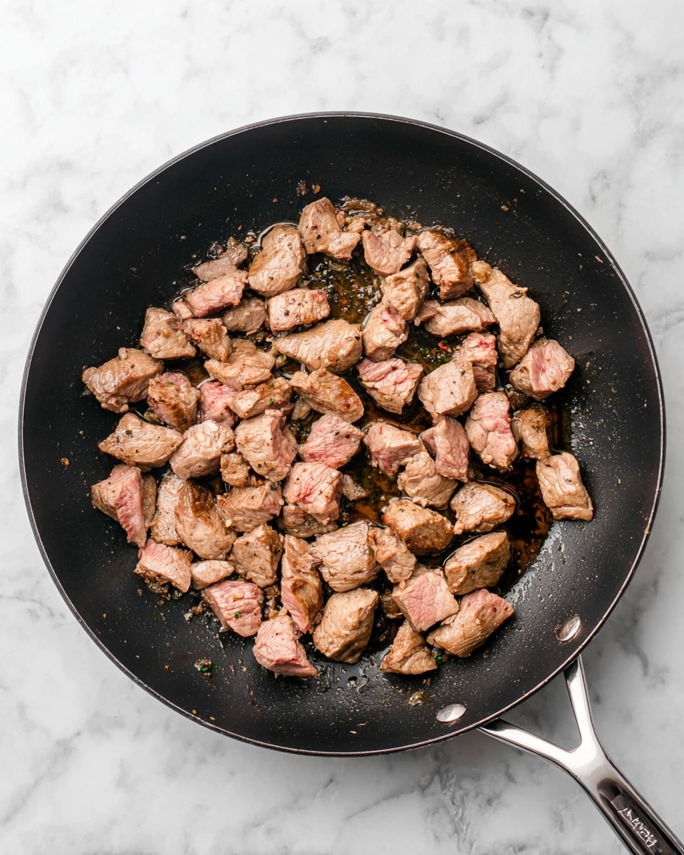 A black frying pan filled with small chunks of cooked meat, showing a mix of light brown and pinkish colors, with some pieces having a slight sear. The pan is on a white marbled surface, and there is a small amount of oil visible around the meat, giving it a shiny texture. The pan's handle is metal and points toward the bottom right corner of the image. Photo taken with an iphone --ar 4:5 --v 7