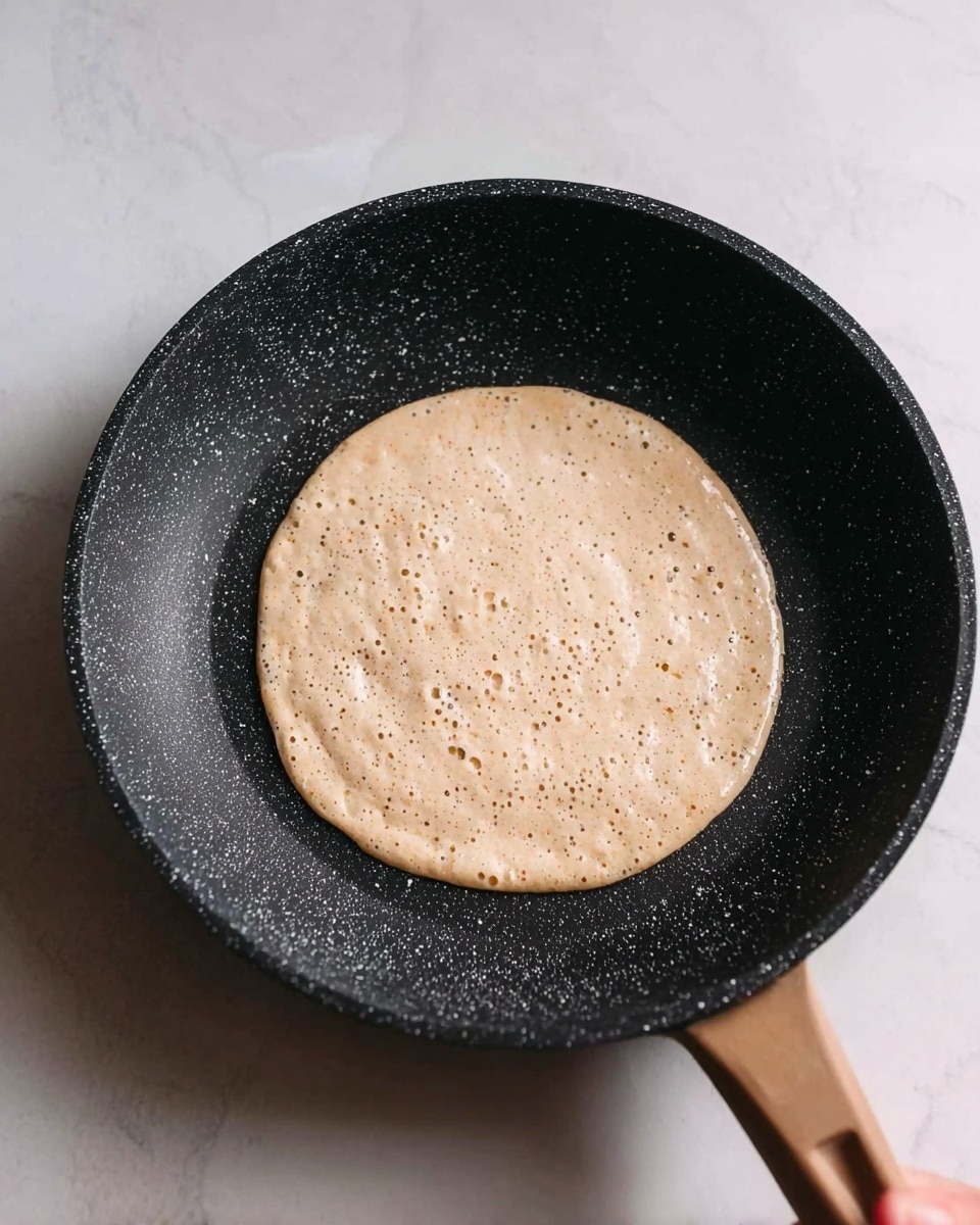 A close-up view of a round pancake batter cooking in a black speckled frying pan, centered in the pan with small bubbles forming on its light beige surface, showing a slightly rough texture. The pan sits on a white marbled surface, with part of a woman's hand visible holding the pan handle on the right edge of the image. Photo taken with an iphone --ar 4:5 --v 7