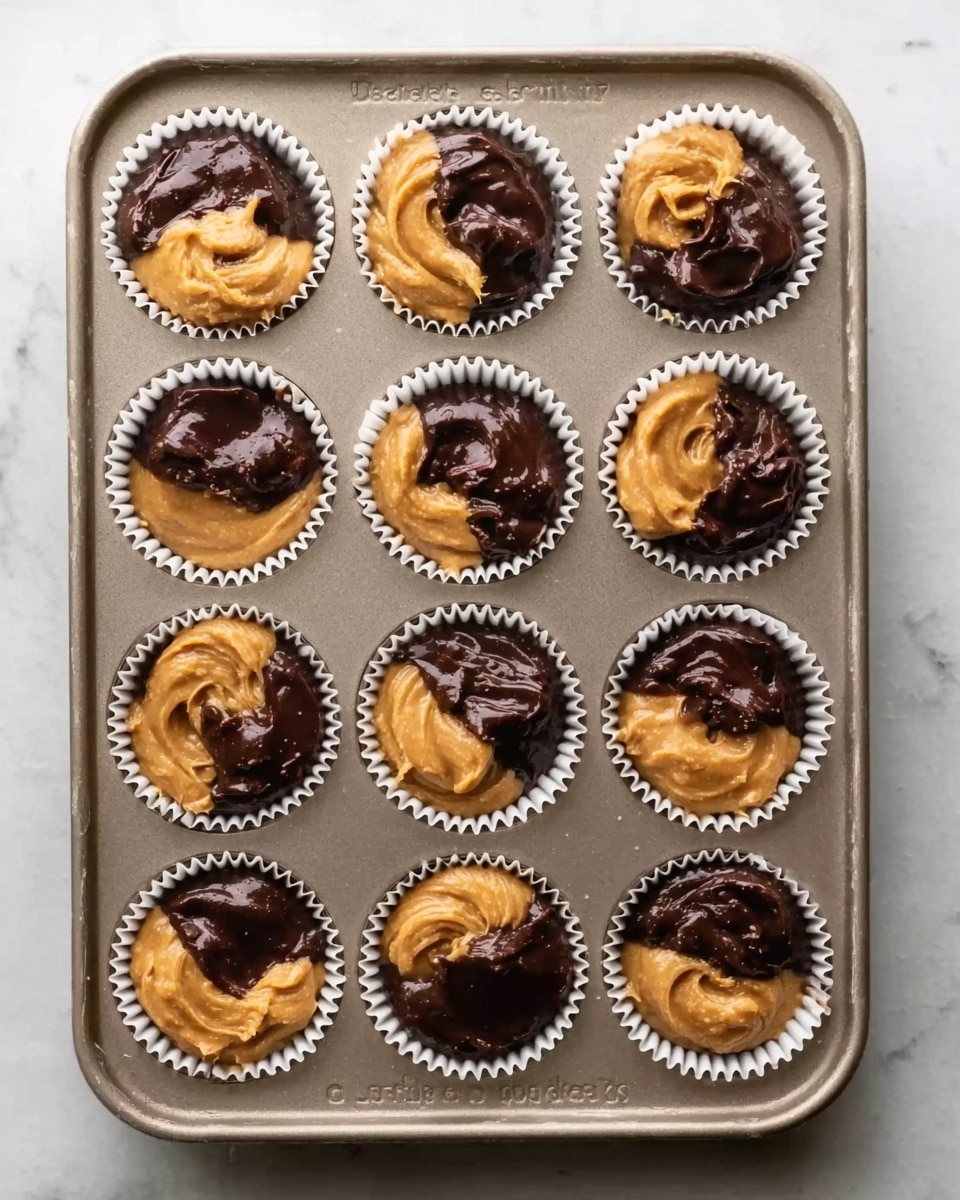 The image shows a metal baking tray filled with twelve white paper cupcake liners. Each liner has two dollops, one of dark brown chocolate batter and one of light golden peanut butter swirled softly side by side, creating a mixed pattern. The chocolate looks smooth and glossy, while the peanut butter has a creamy, thick texture. The tray is placed on a white marbled surface. Photo taken with an iphone --ar 4:5 --v 7