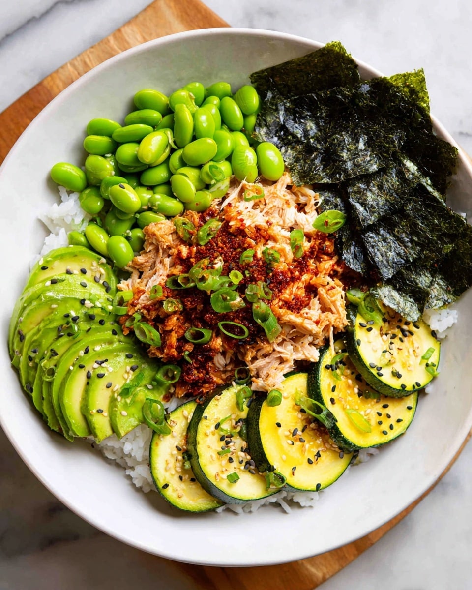 A white bowl on a white marbled surface holds a layer of white rice at the bottom, topped with several layers: bright green edamame beans on one side, shredded light beige chicken in the center sprinkled with dark sesame seeds and a red chili sauce, thin green slices of cucumber with some sauce on the right, and sliced avocado fanned out beside the cucumber. Two pieces of dark green seaweed stick up at the back, adding contrast to the colors and textures of the fresh vegetables and chicken. Photo taken with an iphone --ar 4:5 --v 7