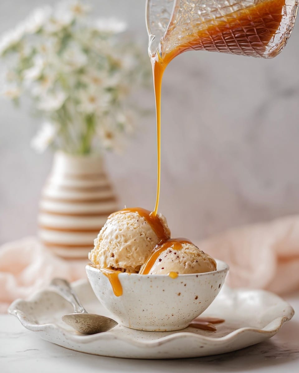 A close-up image of two scoops of light beige ice cream with small dark specks, placed inside a small white speckled bowl which sits in the middle of a white plate with a wavy edge. A smooth caramel sauce is being poured in a thin stream from a textured clear glass container onto the ice cream, with some caramel dripping over the bowl edge and onto the plate. Part of a silver spoon rests on the plate on the left side. The background shows a softly blurred white marbled surface and a striped vase with small white flowers, adding a gentle aesthetic to the scene. Photo taken with an iphone --ar 4:5 --v 7
