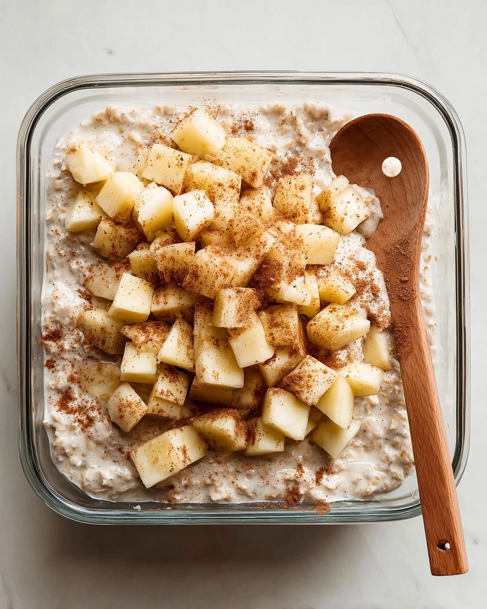 This image shows a piece of apple dessert on a stack of three white plates with a fork resting on the side of the top plate. The dessert has three layers: the bottom layer is a soft, light golden crust; the middle layer is thick with cooked apple slices mixed with creamy filling; the top layer has more apple slices covered with whipped cream and a dusting of cinnamon powder. The plates sit on a blue cloth, and the background has a white marbled texture. There is a white baking dish filled with more apple dessert and some apples blurred in the background. Photo taken with an iphone --ar 4:5 --v 7