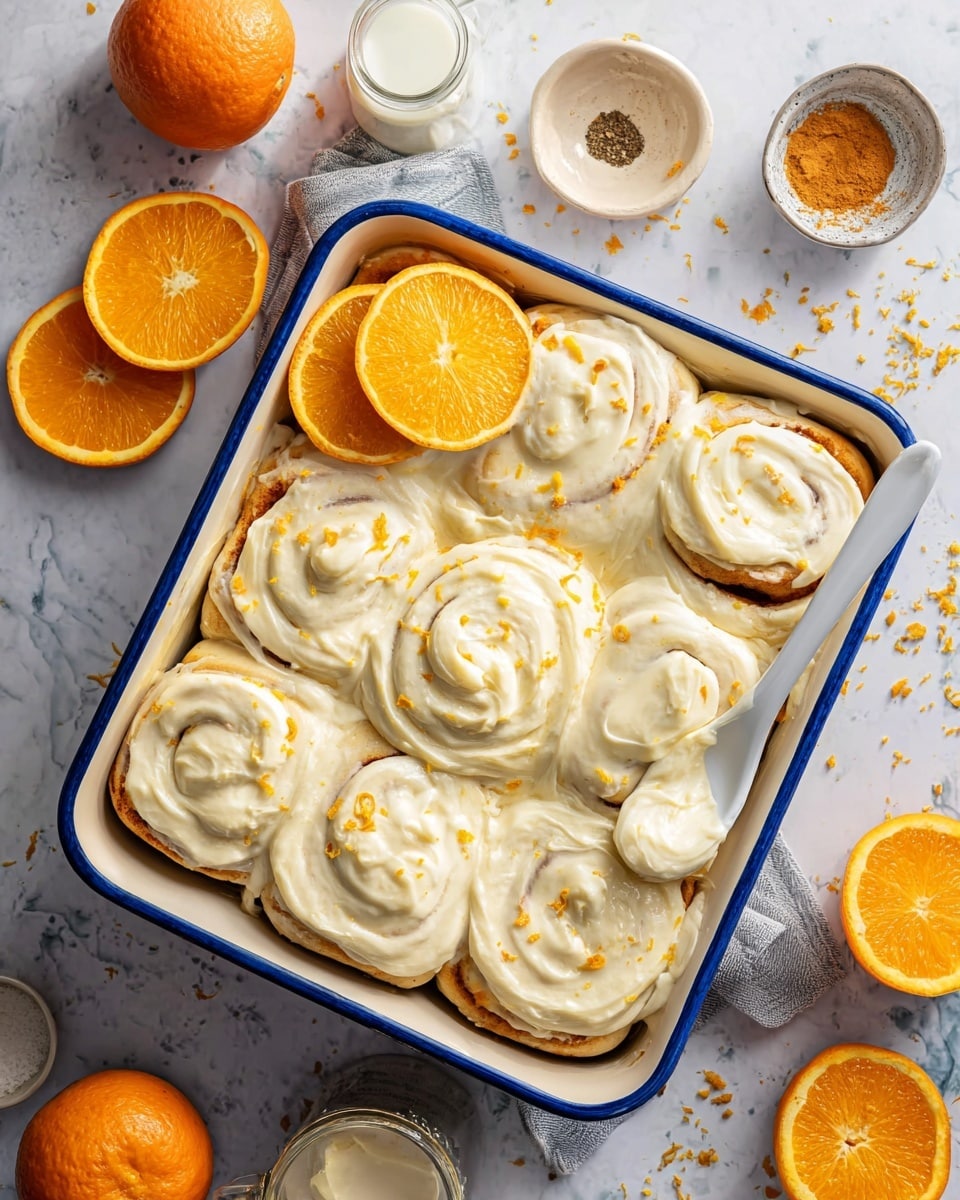 A white square baking dish with a blue rim holds nine cinnamon rolls covered in thick, creamy white frosting that has small orange zest bits sprinkled on top. The cinnamon rolls are arranged in a 3x3 grid, each roll showing a soft, swirled texture under the frosting. In the top left corner of the dish, three thin orange slices lean against one cinnamon roll. A white spoon rests in the middle right roll with frosting clinging to it. The dish is set on a white marbled surface with whole and squeezed oranges scattered around, along with a clear glass of milk and small bowls containing more frosting and ground spices, adding a bright and fresh feel to the scene. Photo taken with an iphone --ar 4:5 --v 7
