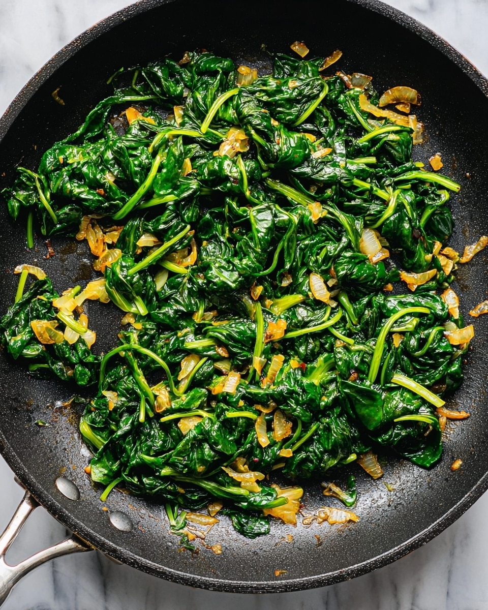 A black frying pan filled with sautéed spinach and diced onions. The spinach is dark green, wilted, and mixed evenly with small, golden-brown pieces of cooked onion, giving a bit of texture contrast. The spinach leaves have visible stems twisted lightly, and the cooking oil adds a slight shine to the overall dish. The pan is placed against a white marbled surface, and the light highlights the fresh look of the greens and the cooked softness of the onions, creating a warm and inviting impression. Photo taken with an iphone --ar 4:5 --v 7