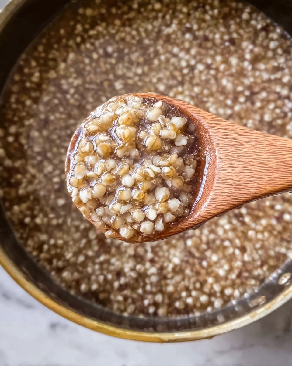 A close-up view of a wooden spoon holding a scoop of cooked buckwheat groats with visible separate grains in light beige and slightly darker shades, surrounded by clear liquid that has a shiny, wet texture. The spoon is above a metal pot filled with more of the same cooked buckwheat and liquid, giving a uniform look of small round grains in light brown and beige tones, with a soft, slightly translucent appearance. The pot edges are round and metallic, providing a subtle contrast to the textured buckwheat inside. The background is a white marbled surface. photo taken with an iphone --ar 4:5 --v 7