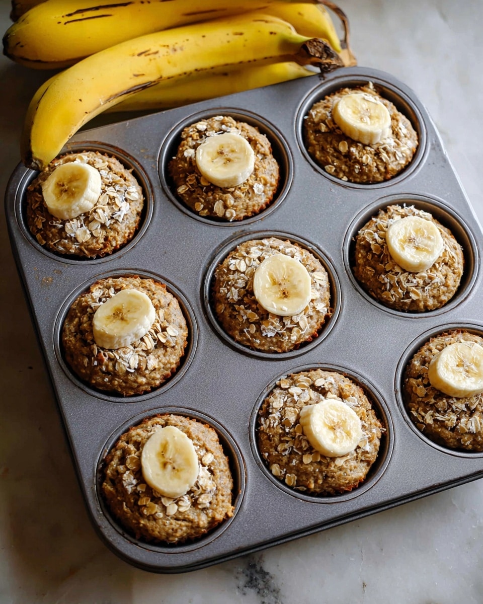 A silver muffin tray holds nine round muffins baking inside, each muffin is topped with a single slightly browned banana slice and sprinkled with tiny oats. The muffins have a coarse, grainy texture and a light brown color with uneven surfaces. Above the tray, a bunch of ripe yellow bananas with a few dark spots is partially visible. The tray is placed on a white marbled surface. photo taken with an iphone --ar 4:5 --v 7