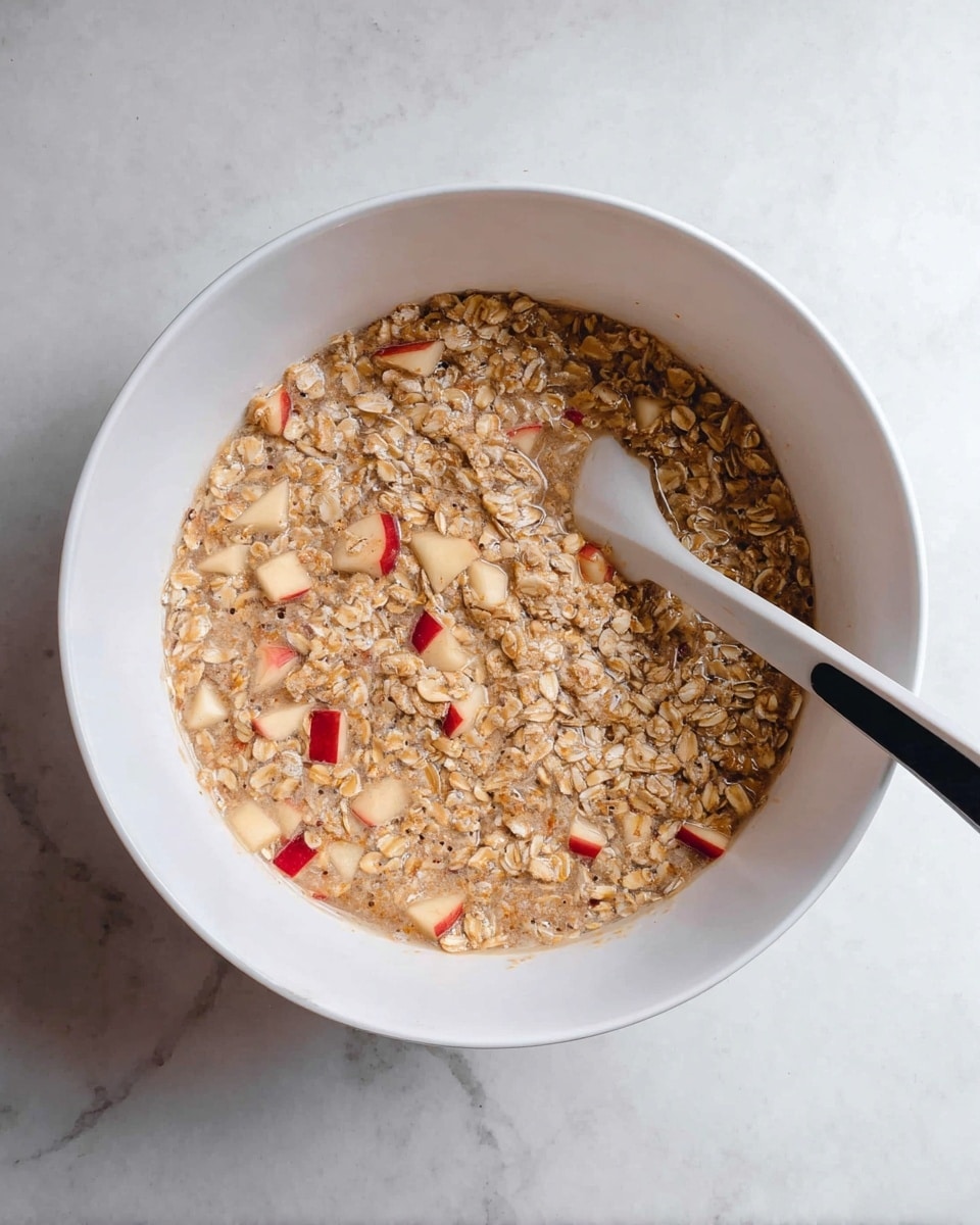 A white bowl filled with a mixture of light brown oats soaked in liquid, dotted with small pieces of red-skinned apple throughout. A white spatula with a black handle rests inside the bowl, partially submerged in the oat mixture. The bowl is placed on a white marbled surface. photo taken with an iphone --ar 4:5 --v 7