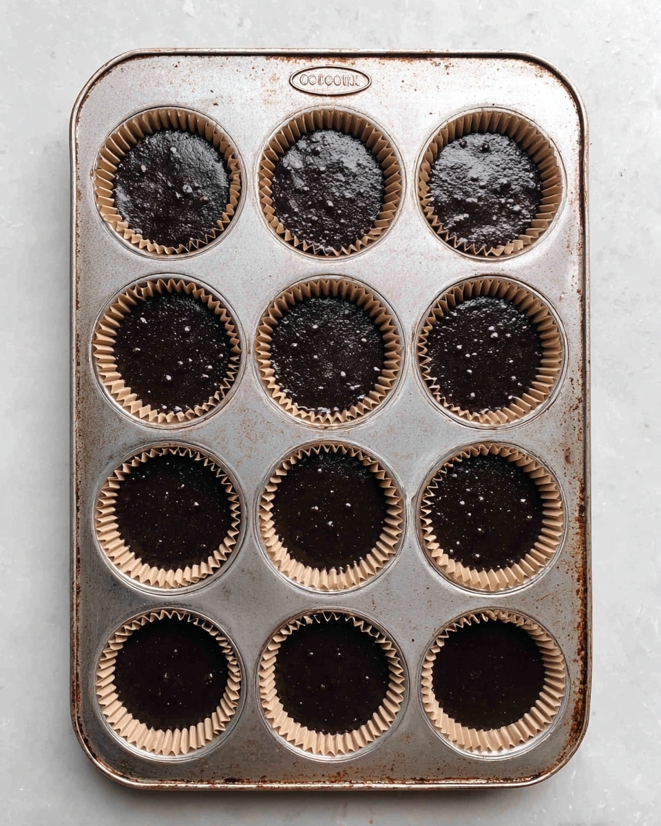 A metal muffin tray filled with 12 brown paper liners, each holding a dark batter that is smooth with small air bubbles on the surface, sitting on a white marbled textured surface. The tray is slightly worn with some rust marks and a logo at the top center. The liners fit neatly inside the round cups of the tray, and the dark batter fills each cup almost to the top, showing a wet, shiny texture. photo taken with an iphone --ar 4:5 --v 7