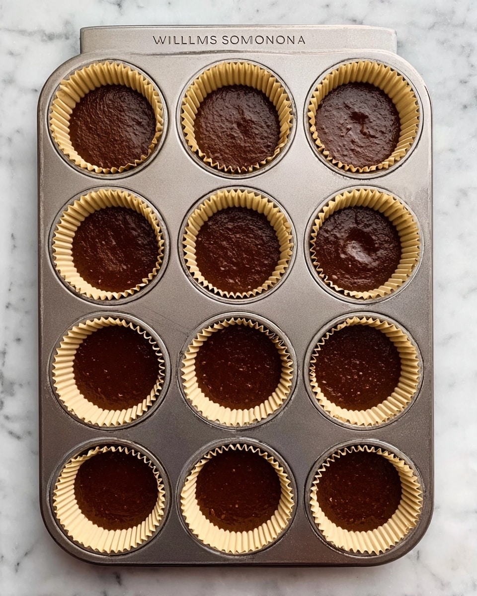 A metal baking tray with twelve round sections, each lined with a light beige paper cup filled with smooth, dark brown batter. The tray is placed on a white marbled surface, and the batter has an even texture with slight bubbles on top, showing it is ready for baking. The tray has the name