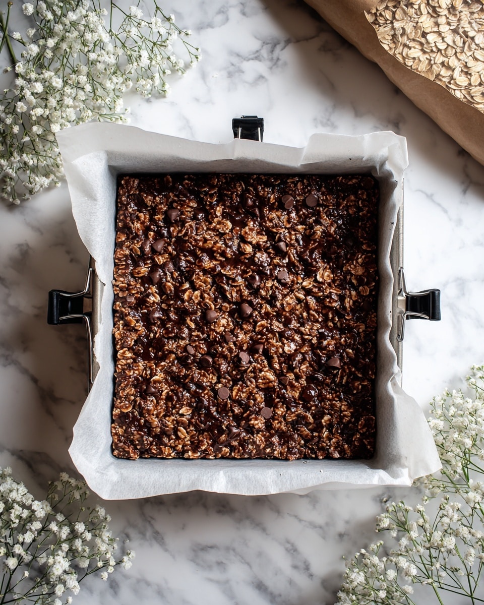 A square baking pan lined with crumpled white parchment paper contains a thick granola bar mixture made of oats and scattered chocolate chips and chunks on its surface. The granola mixture is golden brown with a slight glossy chocolate layer on top, creating a rough texture. The pan is held together by four black binder clips on each corner, placed on a white marbled surface. White baby's breath flowers surround the pan, and a bag of rolled oats with a blue and yellow label is partially visible in the upper right corner. A blush pink card with the number 7 is near the top left corner. photo taken with an iphone --ar 4:5 --v 7
