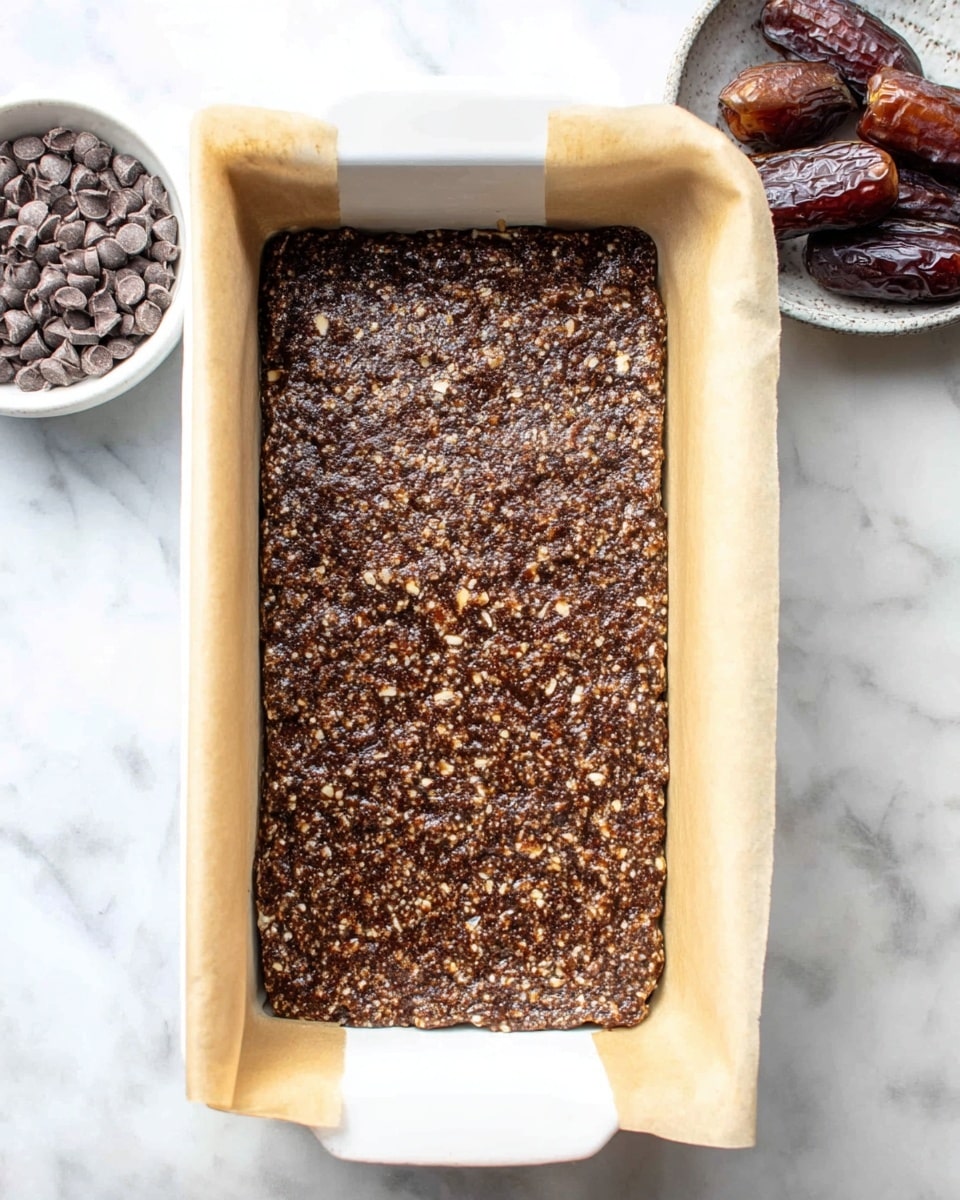 The image shows a rectangular white baking dish lined with parchment paper, inside which there is a thick, evenly spread layer of a dark brown mixture with small visible bits of nuts or seeds. The mixture has a slightly rough texture and fills most of the dish, with the parchment paper edges folded out and resting on the sides. Around the dish, on a white marbled surface, there are two small white bowls, one containing dark dried fruit pieces and the other holding light brown chocolate chips. Photo taken with an iphone --ar 4:5 --v 7
