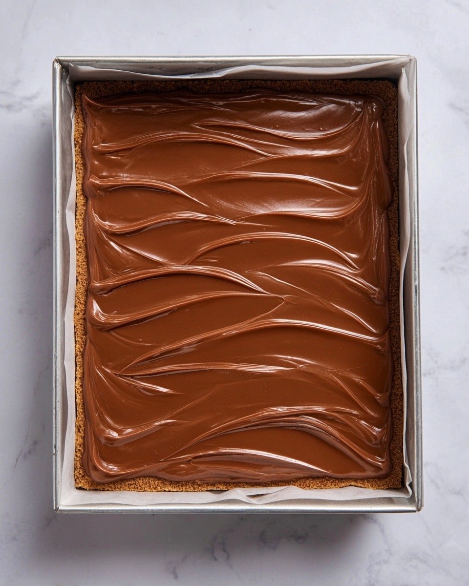 The image shows a square baking pan with three visible layers. The bottom layer is a light brown crumbly crust, topped by a thick, layered filling that looks dense but not very visible from the sides. The top layer is a smooth, shiny milk chocolate spread, evenly covering the entire dessert with gentle, curved swirls running parallel, giving the surface texture. The pan is placed on a white marbled surface with the edges of parchment paper slightly visible inside the pan. photo taken with an iphone --ar 4:5 --v 7