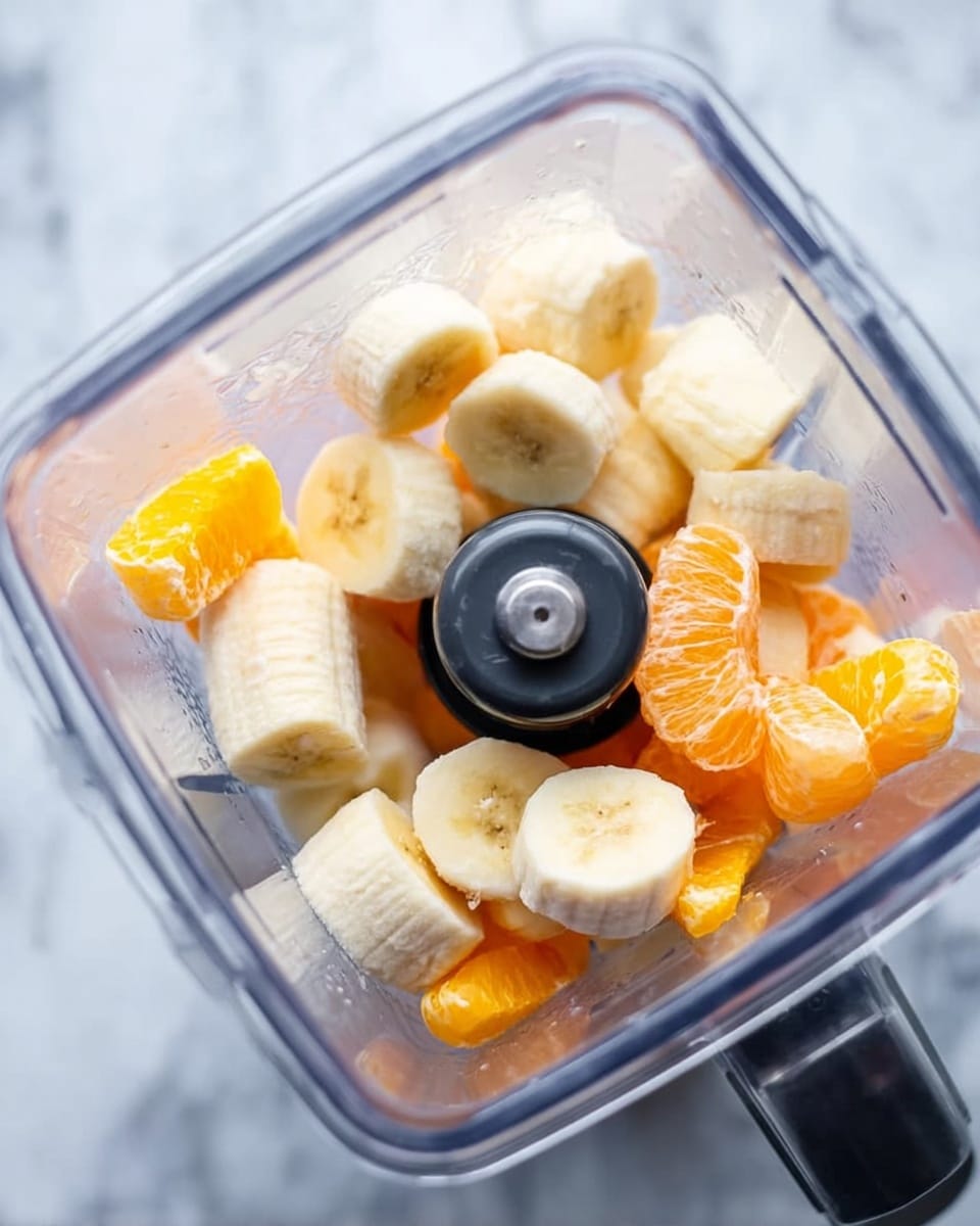 A clear square blender container filled with uneven chunks of light yellow banana slices and bright orange segments, positioned on a white marbled surface. The banana pieces are thick and round, while the orange pieces have a slightly jagged texture and look juicy. The black blender blade and surrounding plastic structure are visible in the center. The image is bright with natural light, enhancing the soft texture of the fruits. Photo taken with an iphone --ar 4:5 --v 7