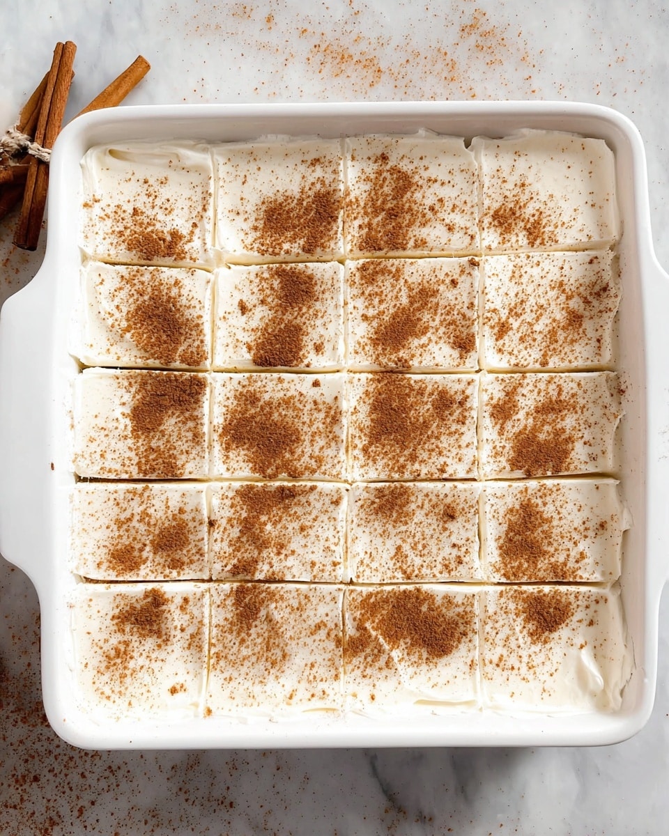 The image shows a white rectangular baking dish filled with a smooth, thick layer of white cream or frosting, sprinkled evenly with a light brown powder, likely cinnamon, covering the whole surface. The dish is divided into 12 rectangular sections by shallow cuts, arranged in three rows and four columns. The white marbled surface beneath the dish adds a clean, bright background. Photo taken with an iphone --ar 4:5 --v 7
