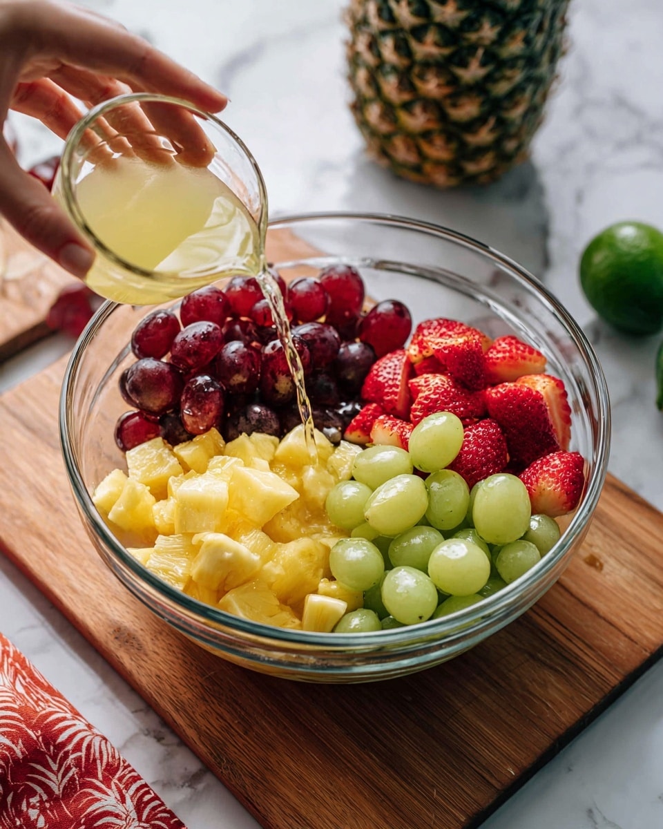 A clear glass bowl filled with four distinct sections of fruit: deep red grapes on the left, bright yellow pineapple chunks in the middle, green grapes at the bottom, and halved red strawberries at the top right. A woman's hand is holding a small clear glass cup, pouring a light yellow liquid over the fruit. The bowl rests on a wooden board, and the background shows a white marbled surface with a green lime half, a whole pineapple top, and a red patterned cloth nearby. photo taken with an iphone --ar 4:5 --v 7