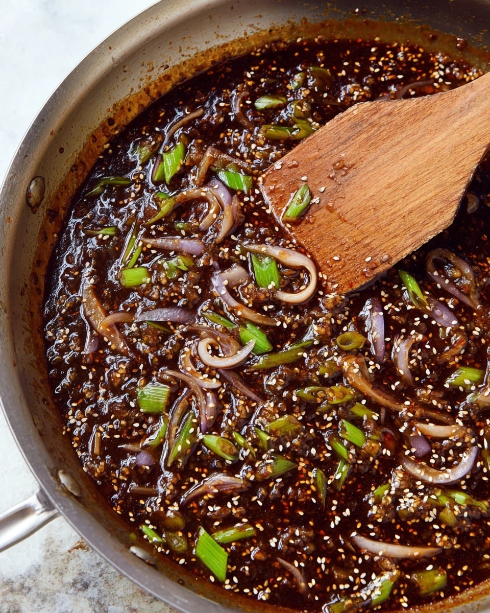 A close-up of a metal pan filled with a thick dark brown sauce mixed with small white sesame seeds and sliced light purple onions. Bright green chopped scallions are scattered throughout the sauce, adding touches of color. A worn, flat wooden spatula is resting inside the sauce on the right side of the pan, slightly lifting some of the mixture. The pan sits on a white marbled texture surface. Photo taken with an iphone --ar 4:5 --v 7