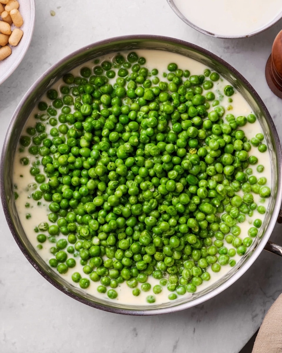 The image shows a shiny metal pan filled with a creamy white sauce as the base layer, topped generously with bright green peas covering almost the entire surface of the sauce. The background is a white marbled texture, and there are small dishes with light brown items partially visible at the top left corner. The scene includes part of a pepper grinder on the right side and a white bowl with a smooth rim at the top edge. Photo taken with an iphone --ar 4:5 --v 7
