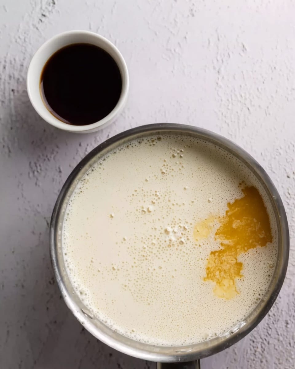 The image shows a metal pot filled with a milky white liquid that has small bubbles and a dollop of golden yellow substance floating on top near the edge. To the top left, there is a small white cup filled with dark brown liquid. The background is a white marbled surface with some subtle gray swirls. Photo taken with an iphone --ar 4:5 --v 7