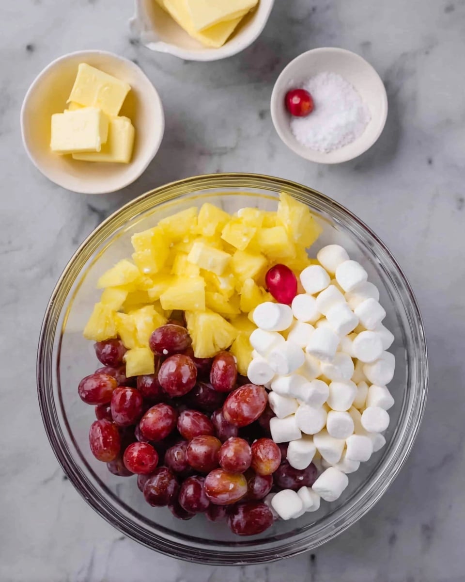A clear glass bowl is placed on a white marbled surface, filled with three layers of ingredients: on the left, bright yellow pineapple chunks; in the middle, red grapes, some cut in half showing their inside; and on the right, a pile of small, white mini marshmallows. Above the bowl, there are two small white bowls; the one on the left holds two small pieces of yellow butter, and the one on the right contains a small amount of white granulated sugar with one red grape. Photo taken with an iphone --ar 4:5 --v 7