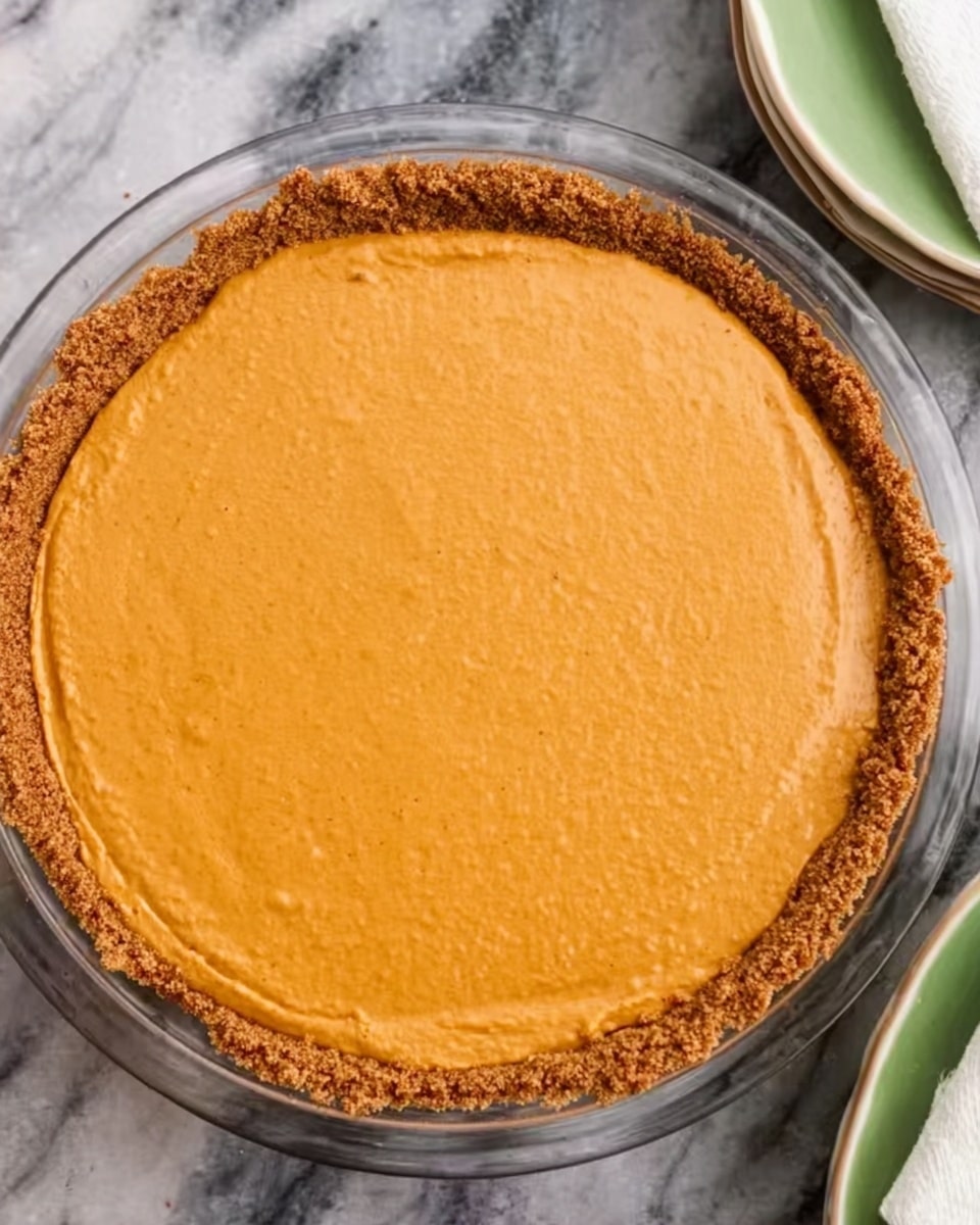 The image shows a round pie with a crumbly brown crust and a smooth, light orange filling on top. The pie is in a clear glass dish, centered on a white marbled surface. The texture of the filling looks creamy and even, covering the entire pie without any decoration. The dish is surrounded by parts of other plates, but the focus is clearly on the pie. Photo taken with an iphone --ar 4:5 --v 7