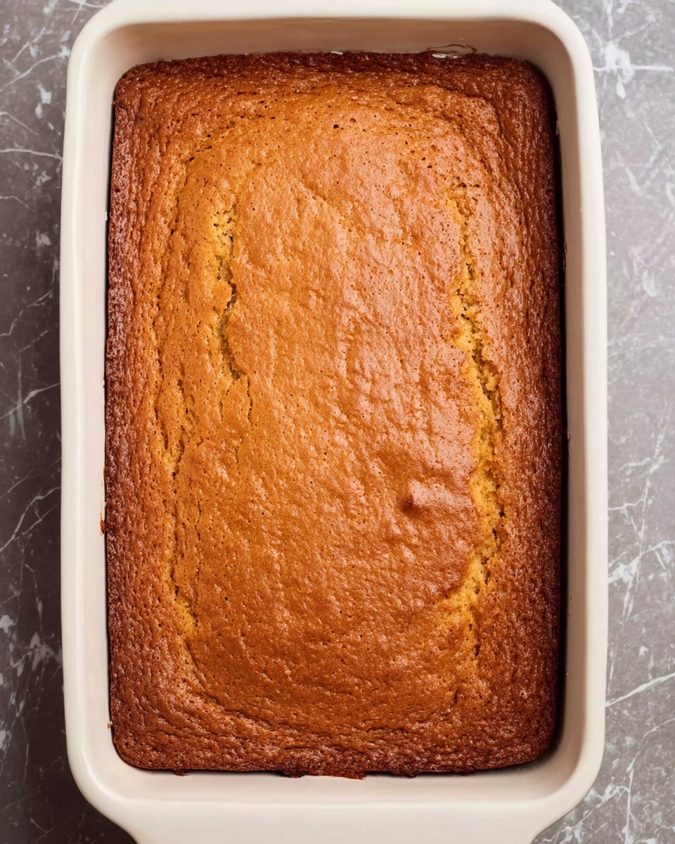 A baked rectangular cake with a golden brown top layer that looks slightly cracked and textured, sitting in a white ceramic baking dish that fills the frame. The edges of the cake are evenly browned. The white marbled surface underneath adds a clean contrast to the warm cake color. The cake appears soft and well-risen with no visible decoration or toppings photo taken with an iphone --ar 4:5 --v 7