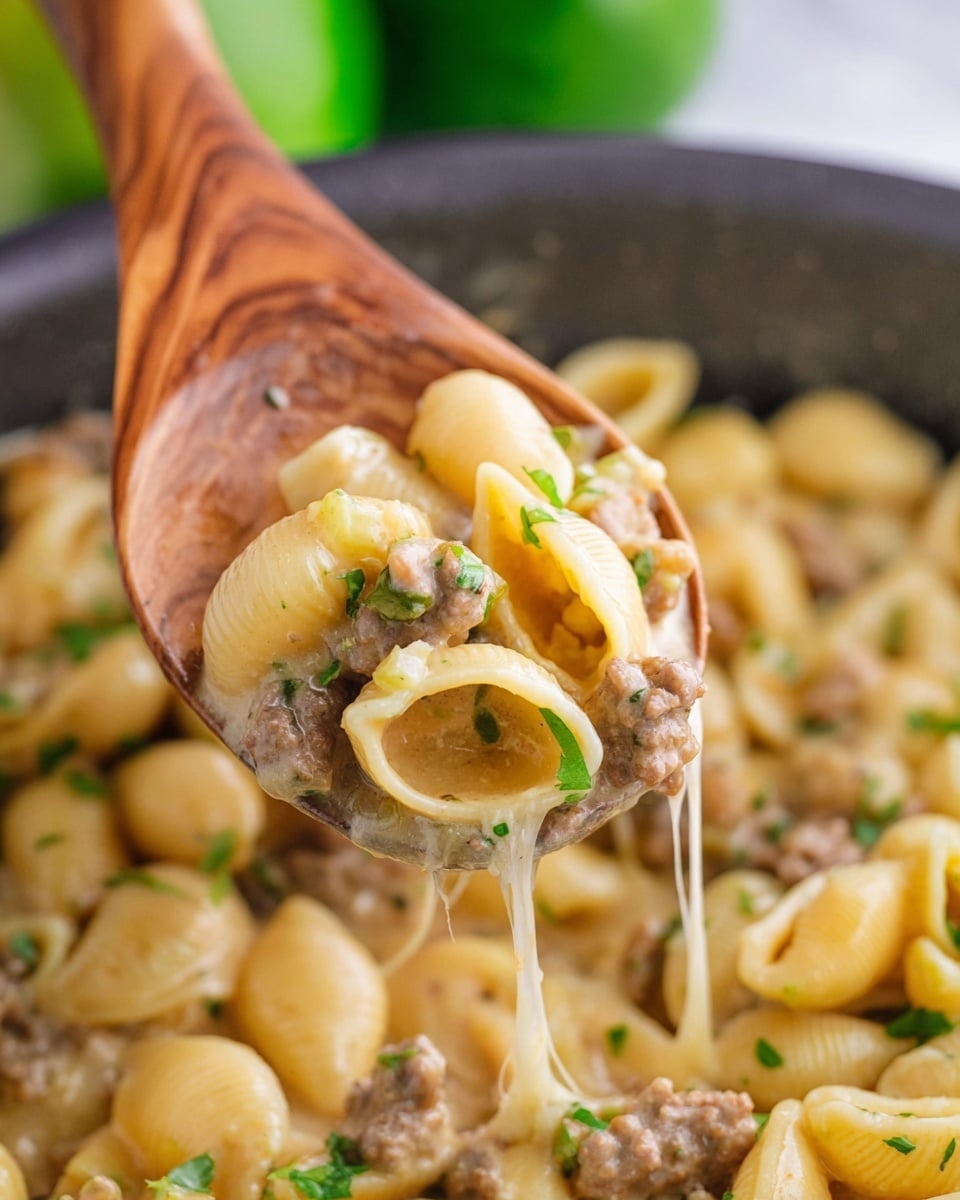 A wooden spoon lifts a scoop of creamy pasta shells mixed with small pieces of ground meat and bits of green herbs, showing a texture of melted cheese stretching below. The pasta is pale yellow with some brown meat pieces and bright green herb leaves scattered on top. The background shows more pasta in a pot with a soft focus and hints of green blurred shapes, all placed against a white marbled surface photo taken with an iphone --ar 4:5 --v 7