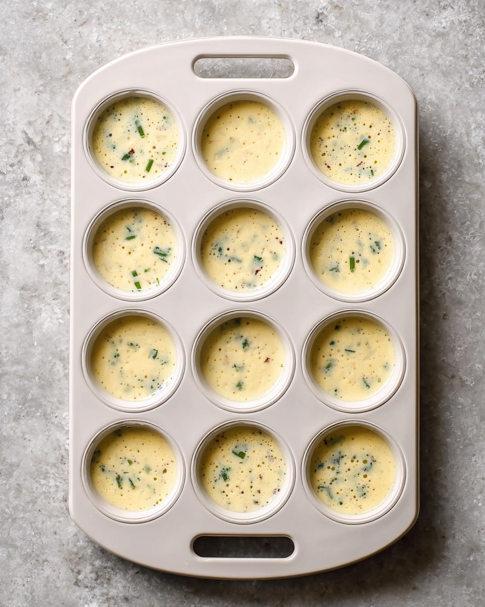 A white tray with twelve round molds is filled with a pale yellow, frothy batter. Small green pieces and some darker bits are spread within the batter in each mold, giving a speckled look. The tray has a handle on the top and bottom, and the batter slightly overflows in some molds. The surface beneath the tray is a white marbled texture. photo taken with an iphone --ar 4:5 --v 7
