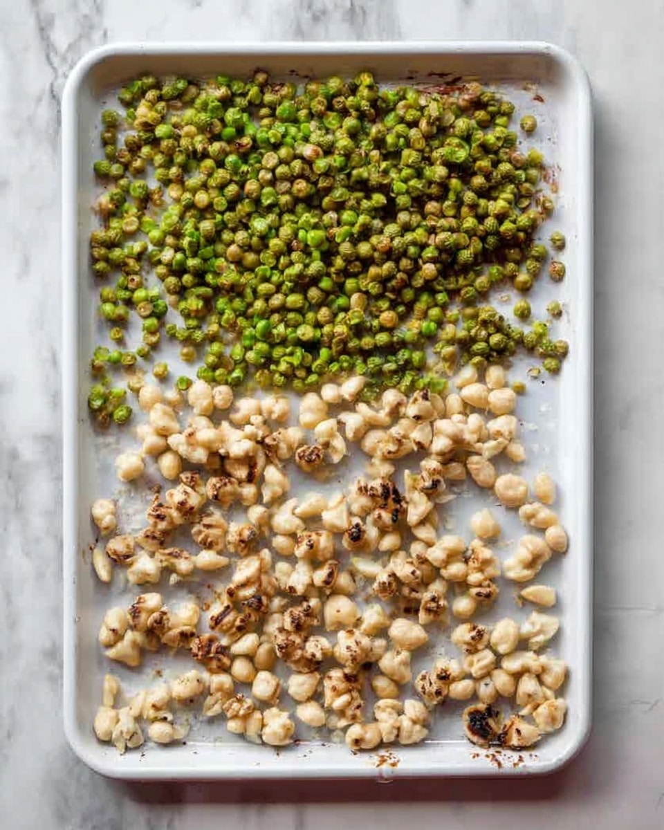 The image shows a white tray on a white marbled surface. On the tray, there are two distinct layers of food. The top layer is made up of small green roasted peas, spread loosely across the top part of the tray. The bottom layer consists of small clusters of light beige cooked nuts, arranged more neatly in rows along the lower part of the tray, with some browning around the edges indicating roasting or cooking. The scene is well-lit and clear, with no other objects visible. Photo taken with an iphone --ar 4:5 --v 7