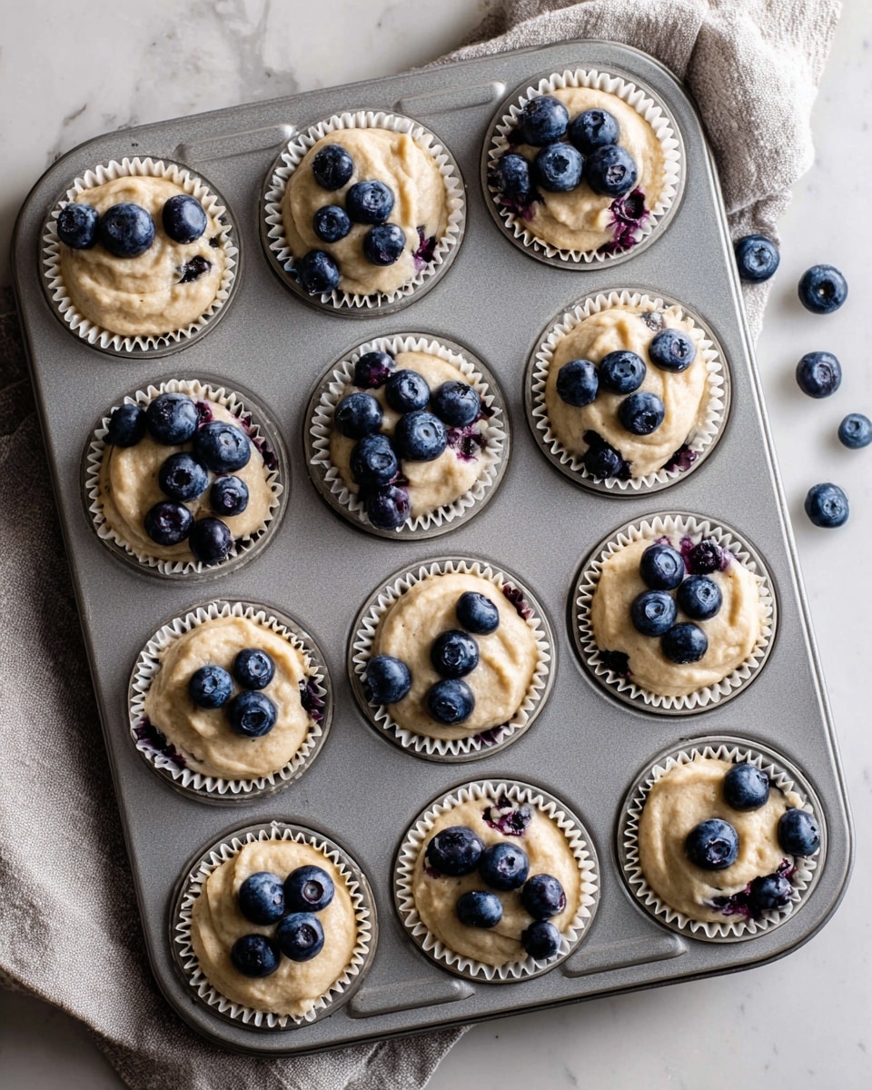 A grey muffin tray holds twelve paper-lined cupcake molds filled with pale tan batter dotted with small dark blueberries. Some molds have more blueberries on top, arranged in small clusters. A woman's left hand holds a small black bowl of blueberries near the tray, while her right hand places blueberries on the batter in one mold. The tray rests on a white marbled surface, creating a clean and bright background. photo taken with an iphone --ar 4:5 --v 7