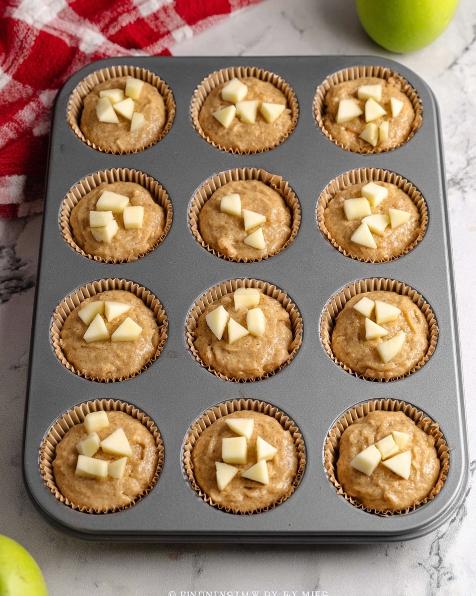 A gray muffin tray holds twelve unbaked muffins lined with light brown paper cups. Each muffin is filled mostly to the top with tan batter that has a slightly lumpy texture, indicating pieces of fruit or nuts inside. On top of each muffin, small, pale yellow chunks are evenly placed, likely pieces of apple. The tray is set on a white marbled surface, with a red and white checkered cloth visible in one corner and a green apple partly in frame nearby. The overall look is neat and ready for baking. photo taken with an iphone --ar 4:5 --v 7