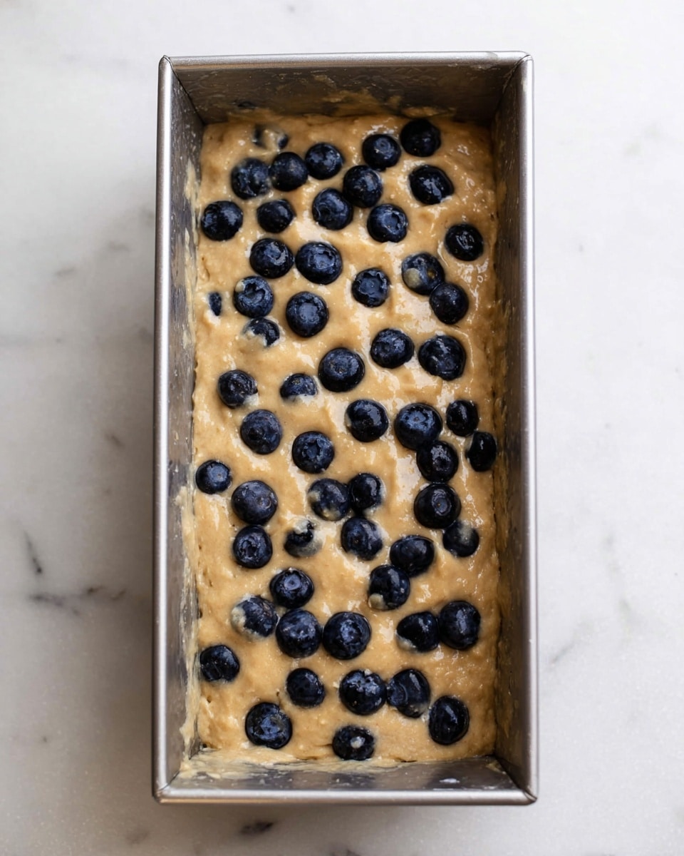 A rectangular silver loaf pan filled with thick light brown batter mixed with whole dark blue blueberries evenly scattered on top. The texture of the batter looks slightly rough and dense. The pan is placed on a white marbled surface. photo taken with an iphone --ar 4:5 --v 7