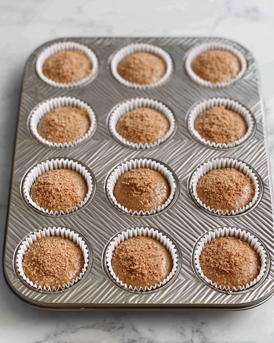 A metal muffin tray with twelve round white paper liners is shown on a white marbled surface. Each liner is filled with brown batter that has a smooth yet slightly textured surface, topped with a dusting of light brown crumbs. The muffin tray has a diagonal striped pattern, and the batter in each cup is level, filling about three-quarters of the liner. The scene is bright with soft natural light. photo taken with an iphone --ar 4:5 --v 7