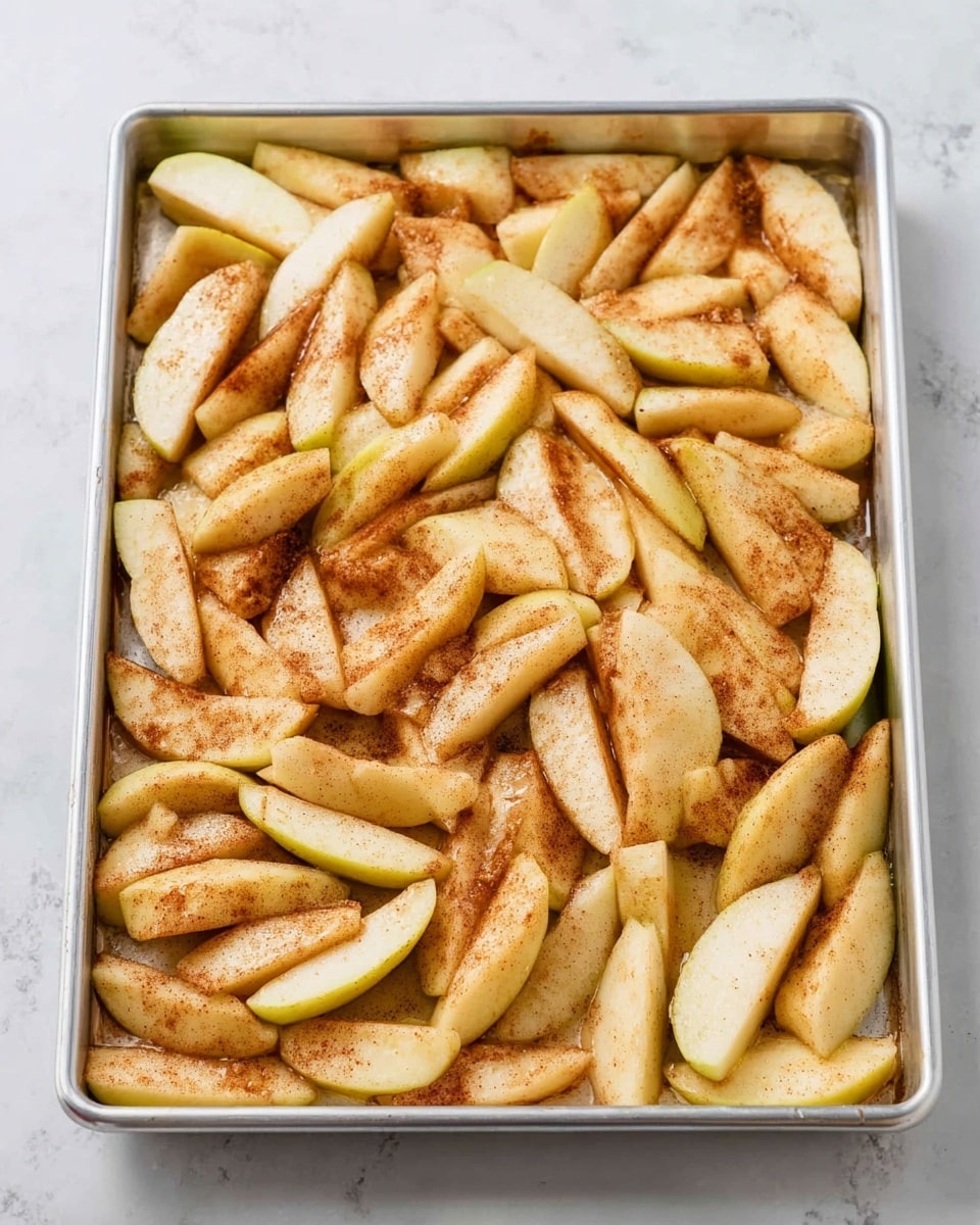 The image shows a large metal tray filled with many slices of apples spread evenly across the surface. The apple slices are pale yellow with a light coating of brown cinnamon or spice dust giving them an uneven golden-brown tint. The slices are soft and slightly shiny, likely due to a light syrup or juice covering them. The tray rests on a white marbled surface. photo taken with an iphone --ar 4:5 --v 7