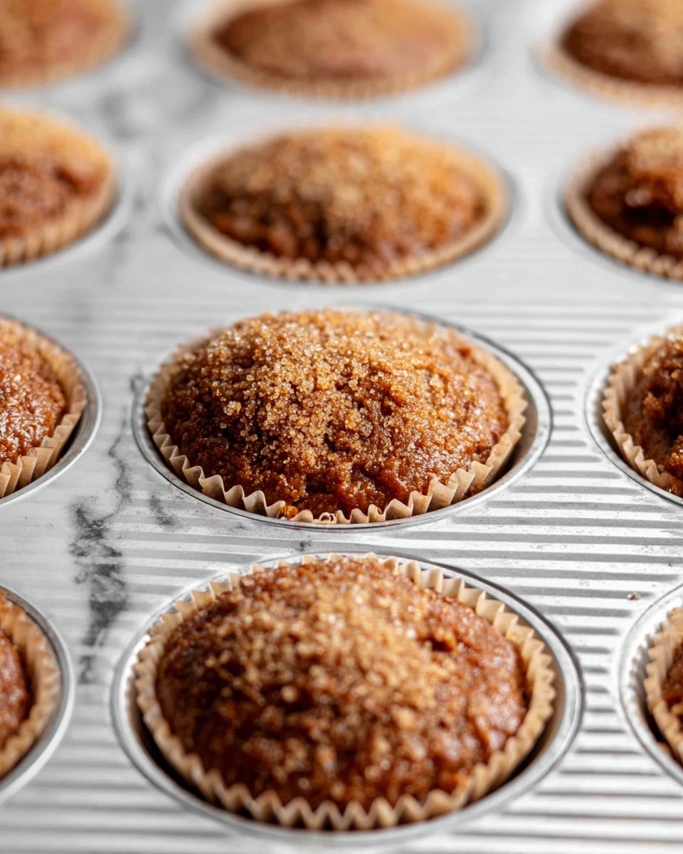 The image shows a close-up of a metal muffin tray with several muffin cups filled with a brown, thick batter that looks like it has small chunks inside. Each muffin cup is lined with light brown paper liners, and the batter is topped with coarse, light tan sugar crystals, giving a rough texture on top. The metal tray has a ribbed surface, and the muffins are slightly rounded on top, showing an uneven, dense texture. The focus is on the middle row, with the rest gently blurred. The background has a white marbled texture. photo taken with an iphone --ar 4:5 --v 7