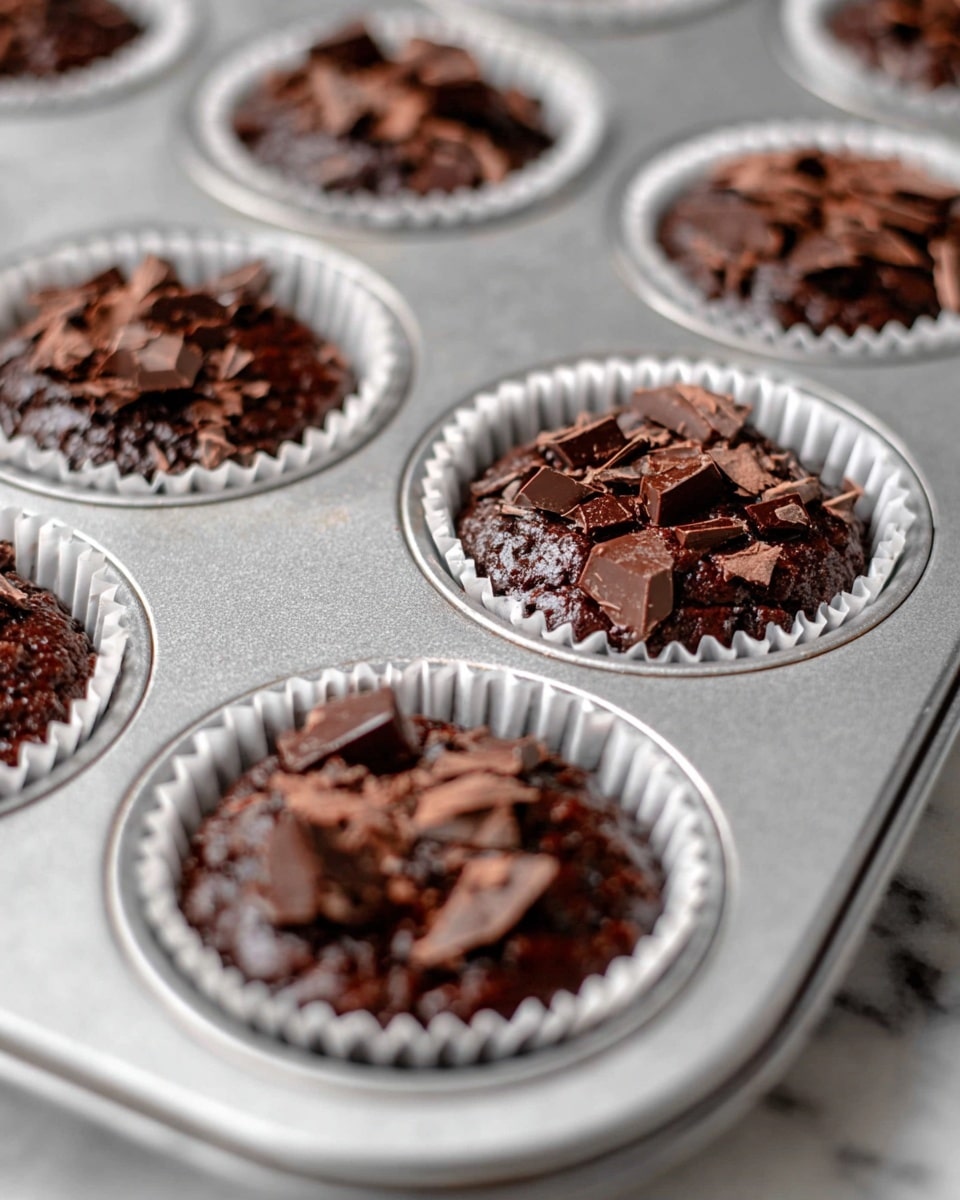 This image shows a metal cupcake tray with white paper liners filled with dark brown chocolate muffin batter. Each muffin is topped with broken pieces of dark chocolate, adding texture and chunks on the glossy, thick batter surface. The tray sits on a white marbled surface. The focus is sharp on the front few muffins, highlighting the rich, dense batter and chocolate pieces, while the background muffins are softly blurred. photo taken with an iphone --ar 4:5 --v 7