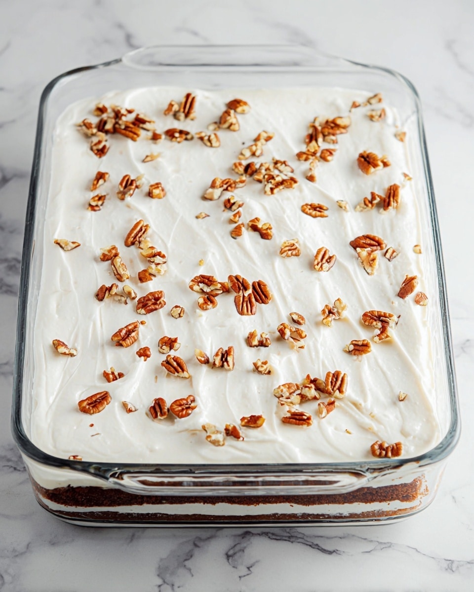 A clear glass rectangular dish placed on a white marbled surface holds a dessert with two visible layers. The bottom layer is dark brown, likely a sticky or moist cake layer. The top layer is thick and white with a smooth, whipped texture that covers the entire dish evenly. Scattered on top are small pieces of toasted pecans, adding a rich brown color and rough texture contrast to the smooth white layer. The dish’s edges are visible, showing how the white layer gently touches the sides. photo taken with an iphone --ar 4:5 --v 7