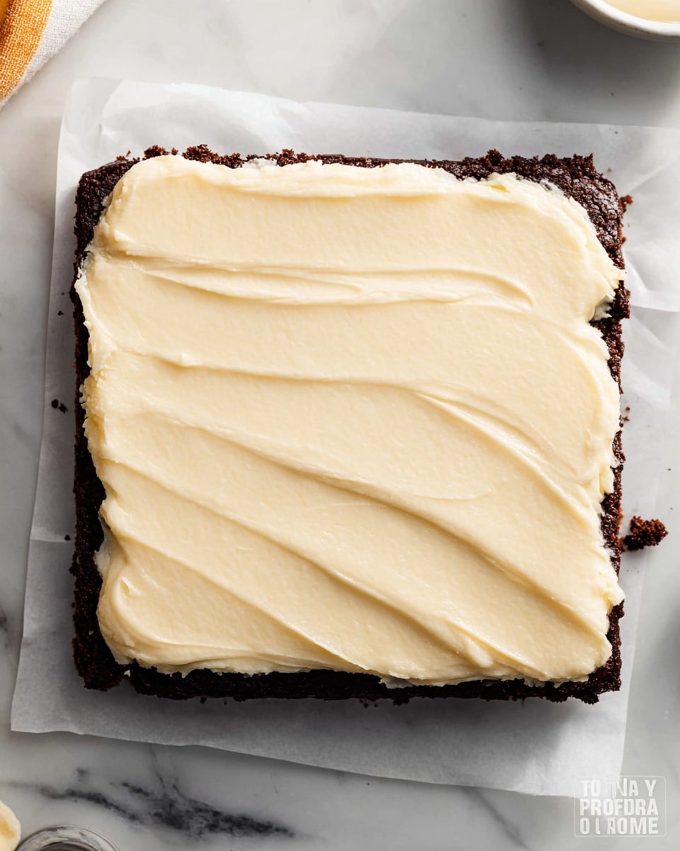 A square dark brown brownie bar is shown from above, topped with a thick, smooth layer of pale cream-colored frosting spread evenly across the entire surface. The brownie base is dense with slightly crumbly edges visible beneath the frosting. The dessert rests on white parchment paper placed on a white marbled surface. A small corner of a white bowl is visible at the edge. Photo taken with an iphone --ar 4:5 --v 7