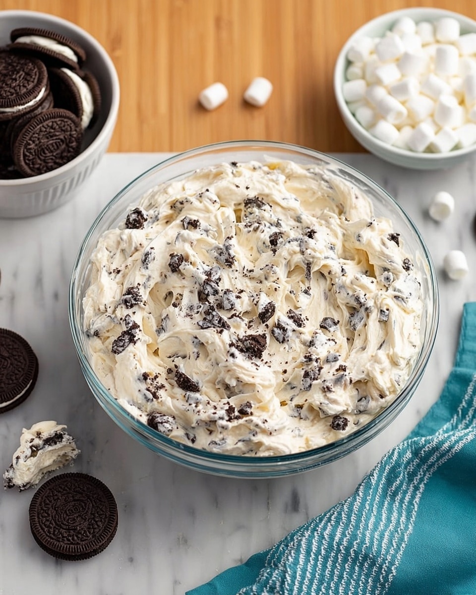 The image shows a clear glass bowl filled with a creamy mixture that is off-white in color with small black cookie pieces mixed throughout, giving it a chunky texture. Surrounding the bowl are a white bowl of whole chocolate sandwich cookies with cream centers, some cookies placed on the white marbled surface with a couple of broken pieces visible, and another white bowl filled with small white marshmallows. A teal and white striped cloth lies beside the cookies on the white marbled surface. The background is a smooth wooden texture. photo taken with an iphone --ar 4:5 --v 7