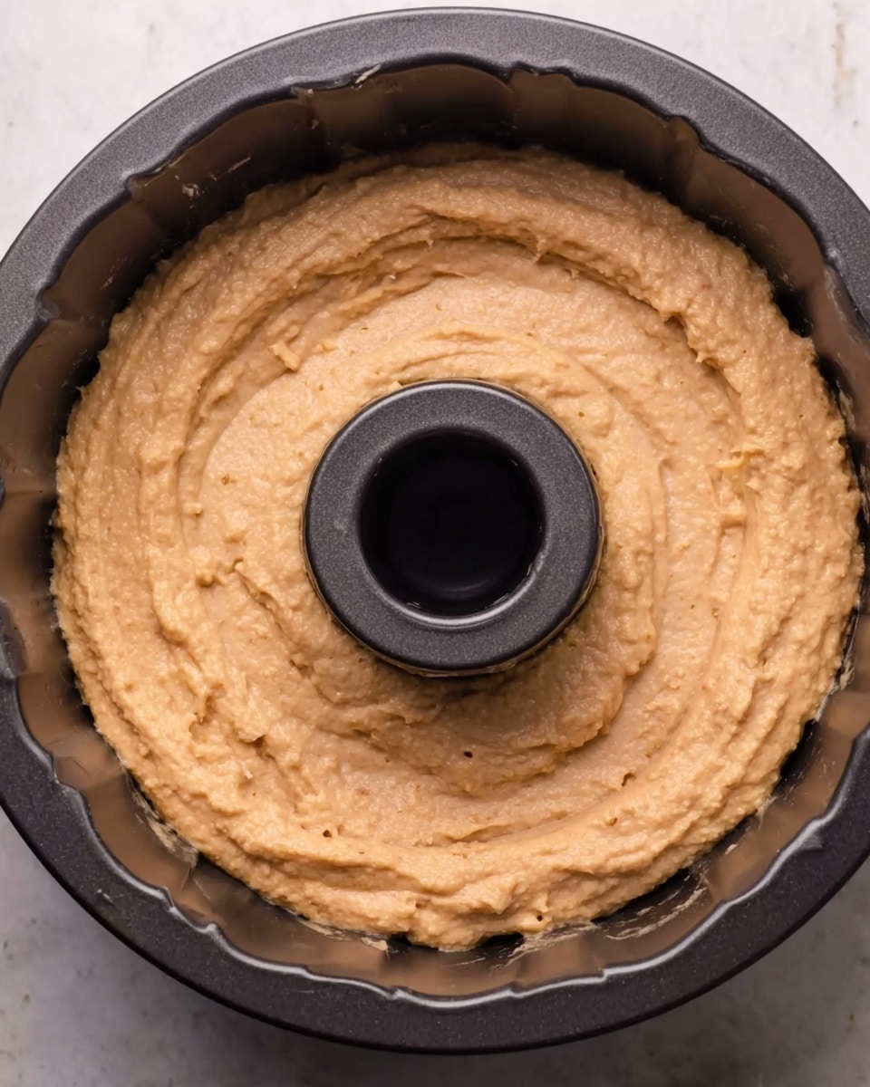 The image shows an unbaked cake batter spread evenly inside a dark, round bundt cake pan with a central hole. The batter is thick and smooth with a light brown color, filling the pan almost fully and touching the inner edges. The surface has a slightly rough texture with small lumps visible. The pan sits on a white marbled surface. Photo taken with an iphone --ar 4:5 --v 7