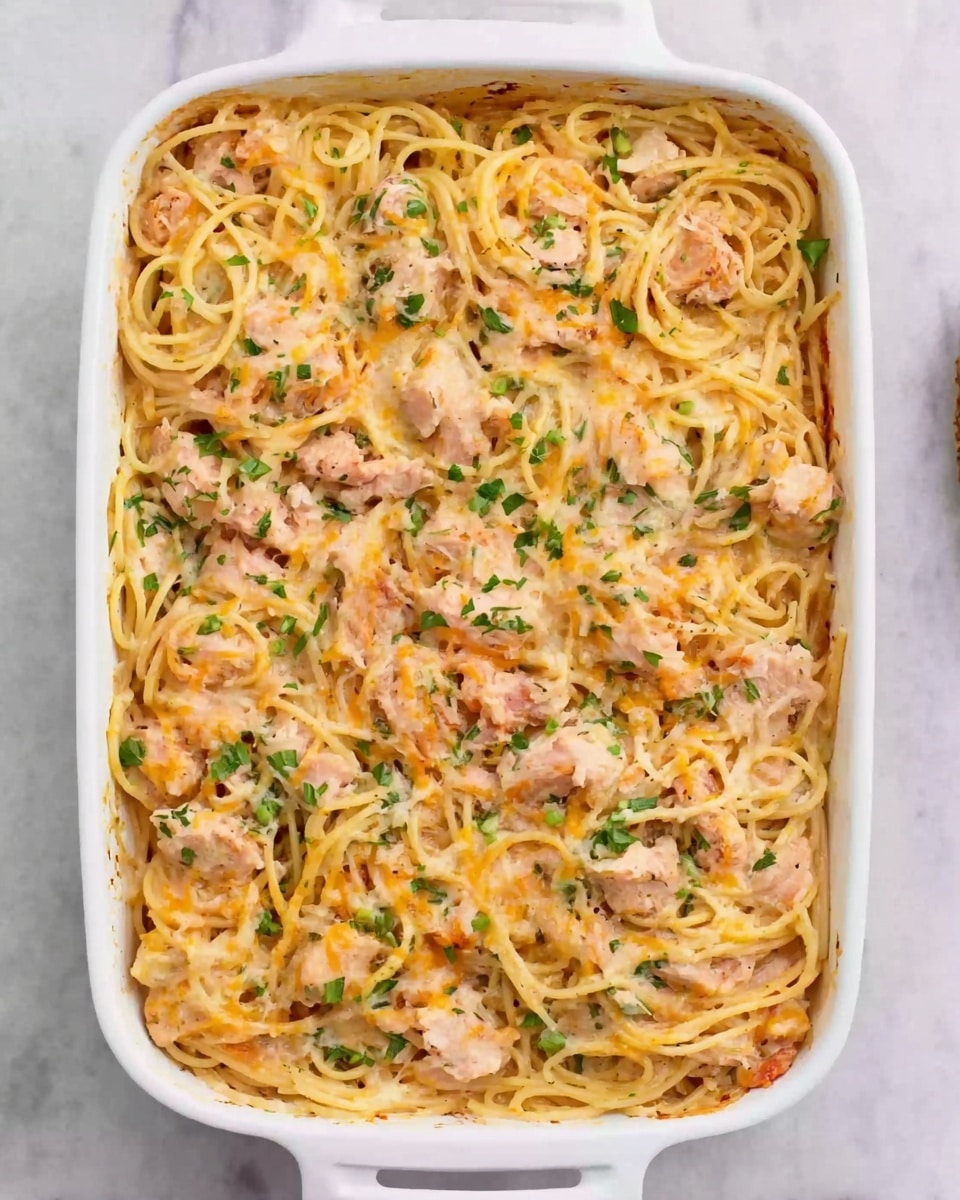 The image shows a white rectangular baking dish filled with a creamy pasta bake. The dish has three visible layers: a base layer of spaghetti coated in a light orange creamy sauce, a middle layer with chunks of light pink chicken mixed with small bits of green herbs and orange cheese, and a top layer where the sauce and cheese are baked slightly golden with green herbs sprinkled on top. The pasta is gently mixed throughout all the layers, with the chicken pieces spread evenly. The background surface is a white marbled texture. Photo taken with an iphone --ar 4:5 --v 7