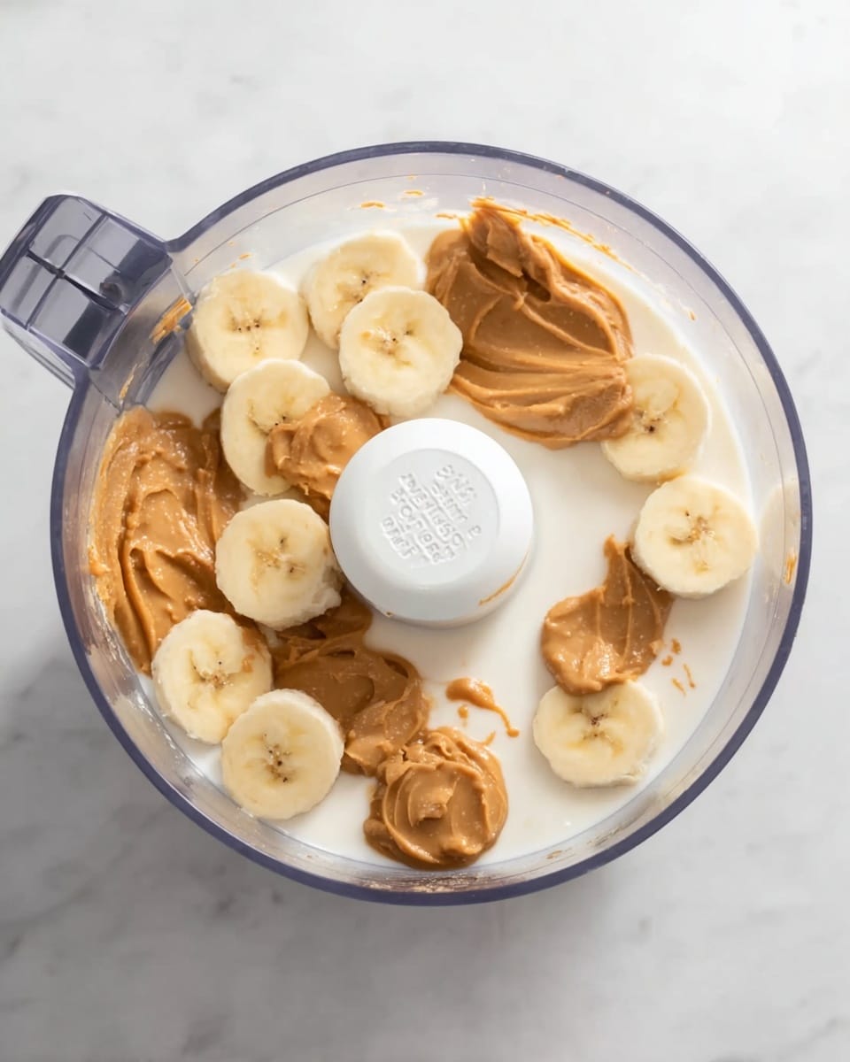 A clear food processor bowl is shown from above, filled with several layers. The bottom layer is a smooth, white milk or cream. On top, there are sliced bananas evenly placed around the inside edge, showing a pale yellow color and soft texture. Dollops of thick, light brown peanut butter are scattered on the banana slices, some larger and some smaller, creating a layered look inside the bowl. The food processor lid has a white central part with text and settings visible. The whole scene sits on a white marbled surface, adding a clean background. Photo taken with an iphone --ar 4:5 --v 7