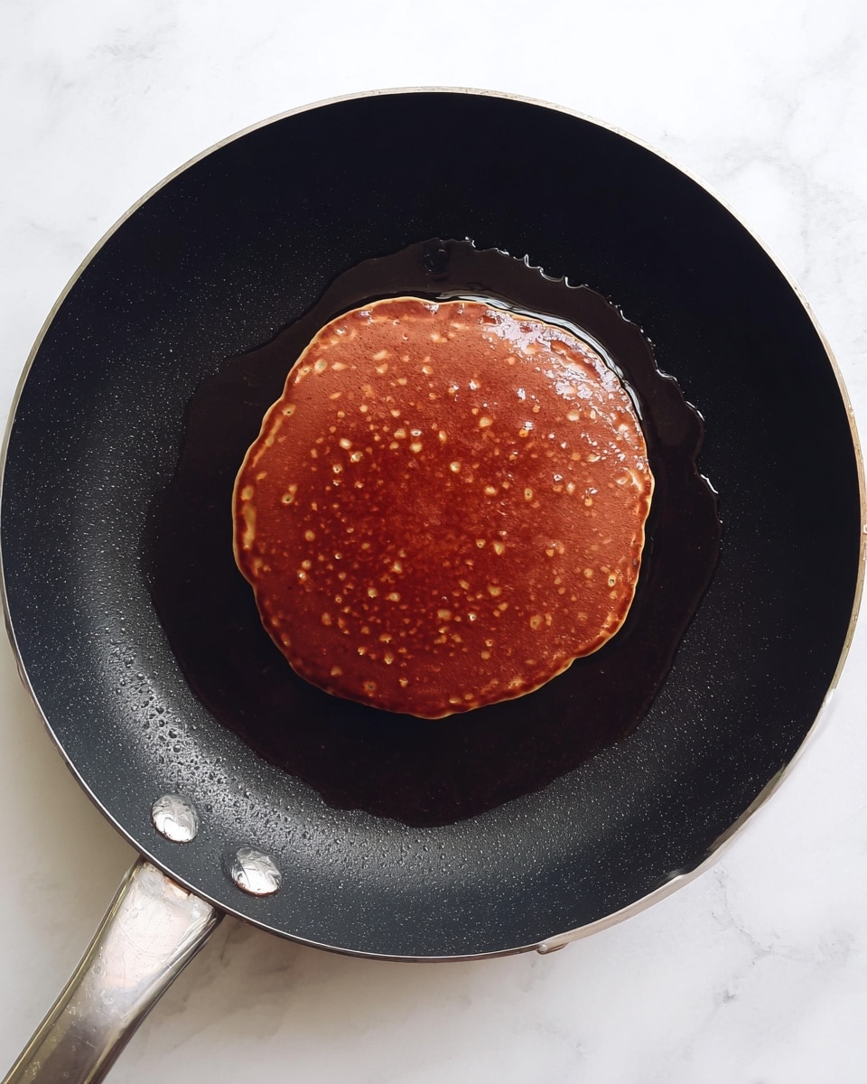 A round pancake cooking in a black frying pan with small bubbles forming on its dark brown surface. The pancake is centered in the pan, which has some shiny melted butter or oil around the edges. The pan's handle is visible at the bottom left, and the background is a white marbled texture. photo taken with an iphone --ar 4:5 --v 7