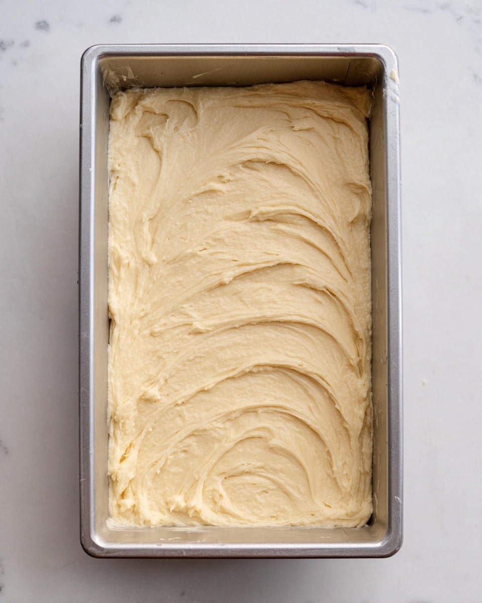 A rectangular silver metal pan filled with one thick layer of light beige batter, spread evenly with visible swirling patterns and a creamy, smooth texture, sitting on a white marbled surface photo taken with an iphone --ar 4:5 --v 7