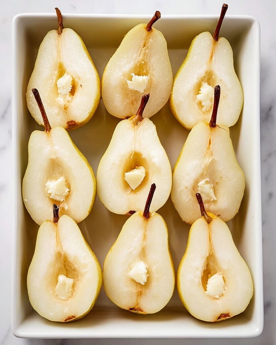 The image shows a white rectangular dish filled with eight halved pears arranged in two rows. Each pear half has a soft light yellowish flesh with a slightly darker center cavity where the seeds were removed. In the middle of each cavity, there is a small dollop of light cream-colored butter-like substance. The pears have their brown stems intact, extending upwards or sideways. The dish is placed on a white marbled surface. photo taken with an iphone --ar 4:5 --v 7