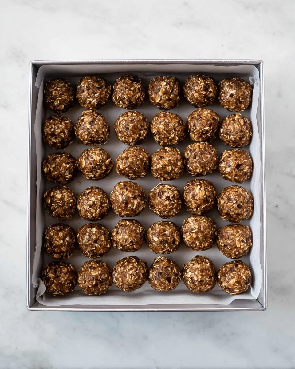 The image shows a silver square pan lined with white parchment paper, filled with 42 small round oat balls arranged neatly in seven rows and six columns. Each oat ball has a rough texture, made of visible oats and small dark spots, giving them a brown, grainy appearance. The pan rests on a white marbled surface, with the oat balls evenly spaced and the lighting soft, highlighting their coarse texture. photo taken with an iphone --ar 4:5 --v 7
