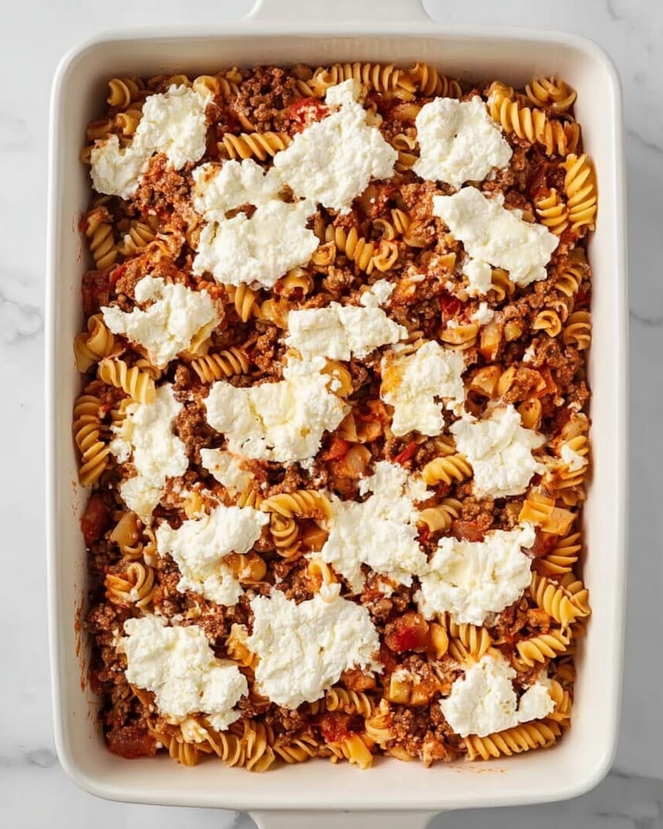 A white rectangular baking dish is filled with one layer of cooked pasta mixed with ground meat and small bits of vegetables in a reddish sauce. On top, there are dollops of a white creamy mixture, likely a cheese blend, spread unevenly over the pasta. The dish sits on a white marbled surface, showing a clear contrast between the warm colors of the pasta and meat and the cool, smooth white of the cheese topping. Photo taken with an iphone --ar 4:5 --v 7