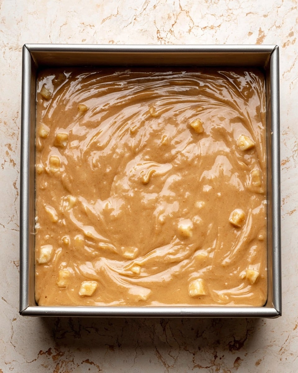 A square metal baking pan filled with light brown batter that has a smooth, thick texture with small chunks of pale yellow fruit mixed throughout. The batter surface shows gentle swirls and glossiness, indicating it is freshly poured and not yet baked. The pan sits on a white marbled textured surface. photo taken with an iphone --ar 4:5 --v 7
